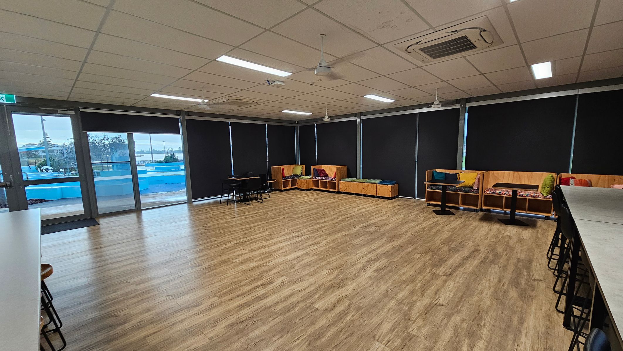 A spacious room with wooden flooring and a ceiling with fluorescent lights and air conditioning units. On the left, there are large windows with black blinds and a view of a blue outdoor area. In the centre, there are a few tables and chairs. On the right, there are wooden benches with colourful cushions. A long counter with high stools is also visible on the right side. A spacious room with wooden flooring and a ceiling with fluorescent lights and air conditioning units. On the left, there are large windows with black blinds and a view of a blue outdoor area. In the centre, there are a few tables and chairs. On the right, there are wooden benches with colourful cushions. A long counter with high stools is also visible on the right side.