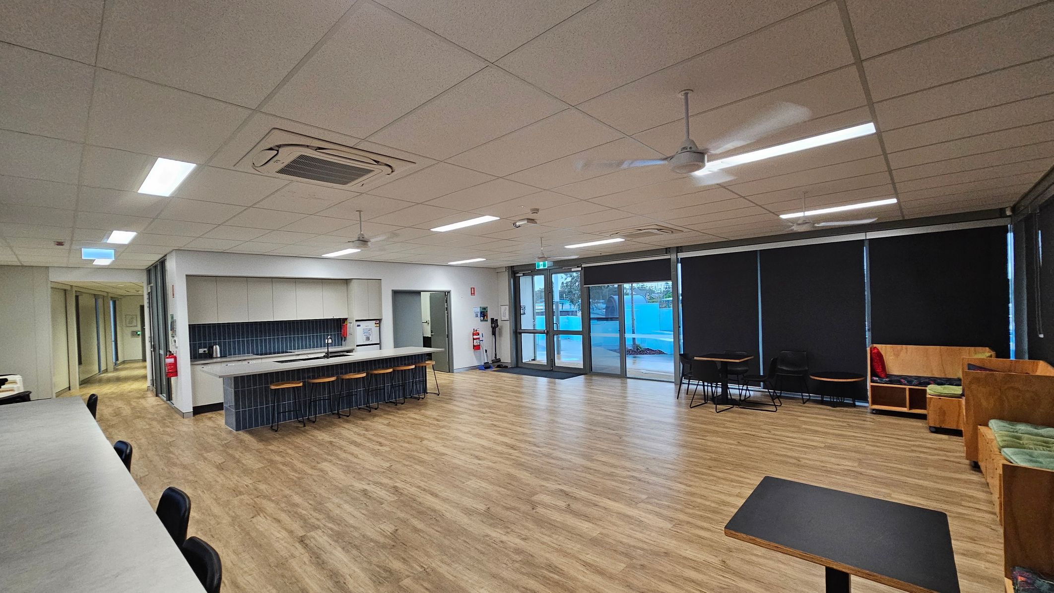 A spacious room with a light wooden floor and a tiled ceiling. On the left, there's a long counter with black chairs. In the middle, a kitchen area features a counter with stools, a sink, and cabinets. The right side has large windows with black blinds, a few tables, and wooden benches with cushions. Ceiling fans and lights are evenly spaced across the ceiling. A corridor is visible on the far left. A spacious room with a light wooden floor and a tiled ceiling. On the left, there's a long counter with black chairs. In the middle, a kitchen area features a counter with stools, a sink, and cabinets. The right side has large windows with black blinds, a few tables, and wooden benches with cushions. Ceiling fans and lights are evenly spaced across the ceiling. A corridor is visible on the far left.