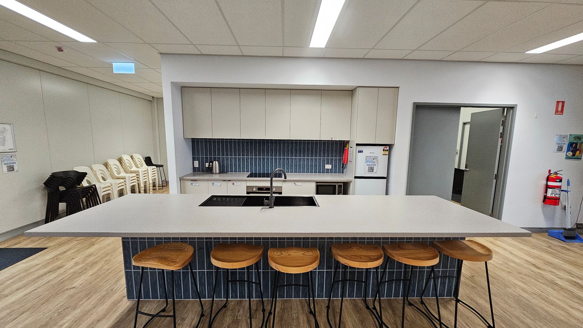 A modern kitchen area with a large island in the centre featuring a white countertop and a built-in sink. Five wooden bar stools with metal legs are lined up in front. The back wall has light-coloured cabinets and a blue-tiled backsplash. Appliances include a kettle, stovetop, and a fridge. To the left, stacked white and black chairs are against the wall. On the right, a fire extinguisher is mounted near an open door. The floor is wooden, and the ceiling has recessed lighting. A modern kitchen area with a large island in the centre featuring a white countertop and a built-in sink. Five wooden bar stools with metal legs are lined up in front. The back wall has light-coloured cabinets and a blue-tiled backsplash. Appliances include a kettle, stovetop, and a fridge. To the left, stacked white and black chairs are against the wall. On the right, a fire extinguisher is mounted near an open door. The floor is wooden, and the ceiling has recessed lighting.
