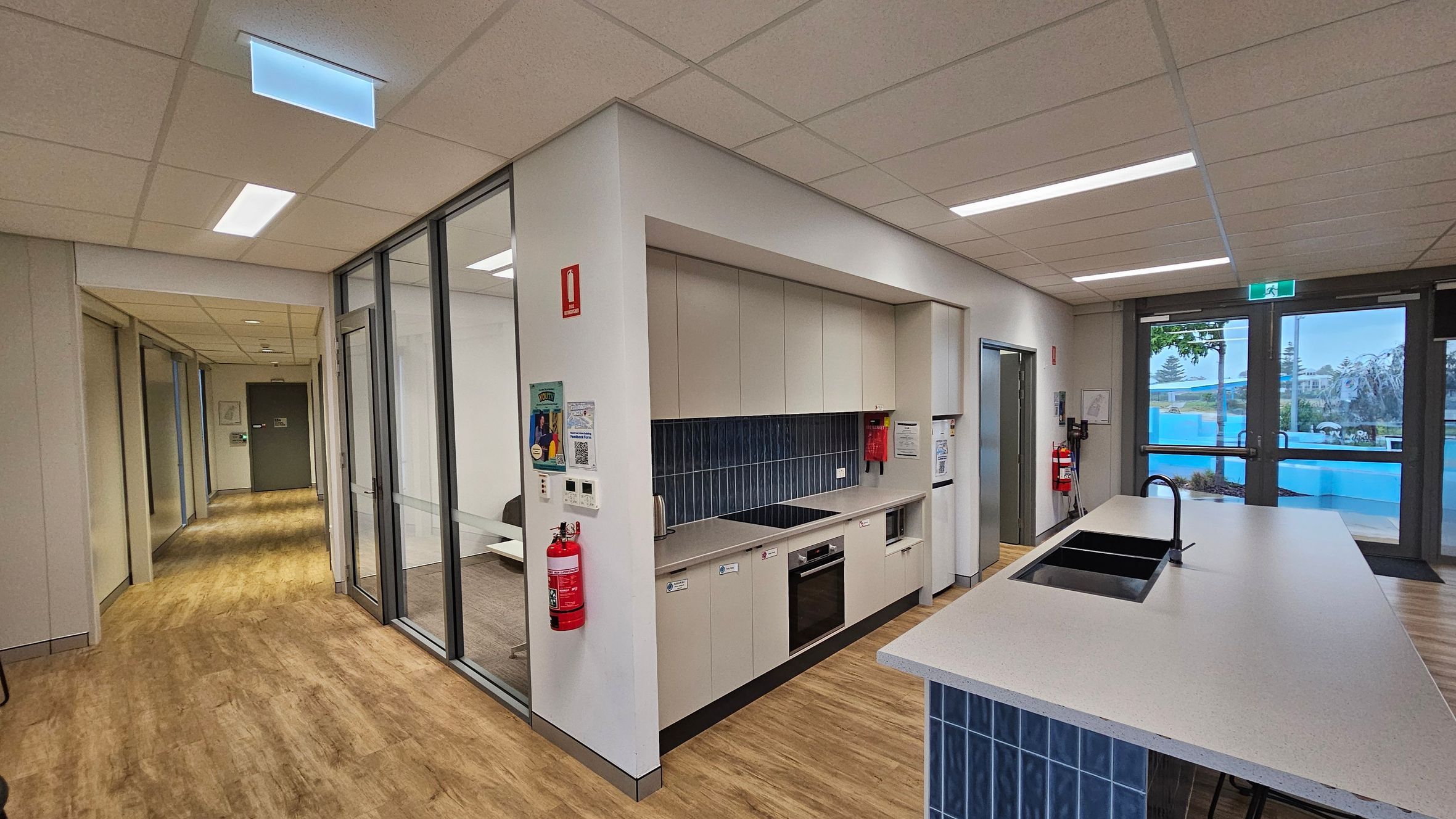 A modern kitchen area with light wood flooring and a suspended ceiling with recessed lighting. On the left, there is a hallway leading to a green door. In the centre, a kitchenette with beige cabinets, a black cooktop, and a built-in oven. A fire extinguisher is mounted on the wall. On the right, a large island with a sink and tap, and glass doors leading outside. Blue tiles are used as a backsplash and on the island base. A modern kitchen area with light wood flooring and a suspended ceiling with recessed lighting. On the left, there is a hallway leading to a green door. In the centre, a kitchenette with beige cabinets, a black cooktop, and a built-in oven. A fire extinguisher is mounted on the wall. On the right, a large island with a sink and tap, and glass doors leading outside. Blue tiles are used as a backsplash and on the island base.