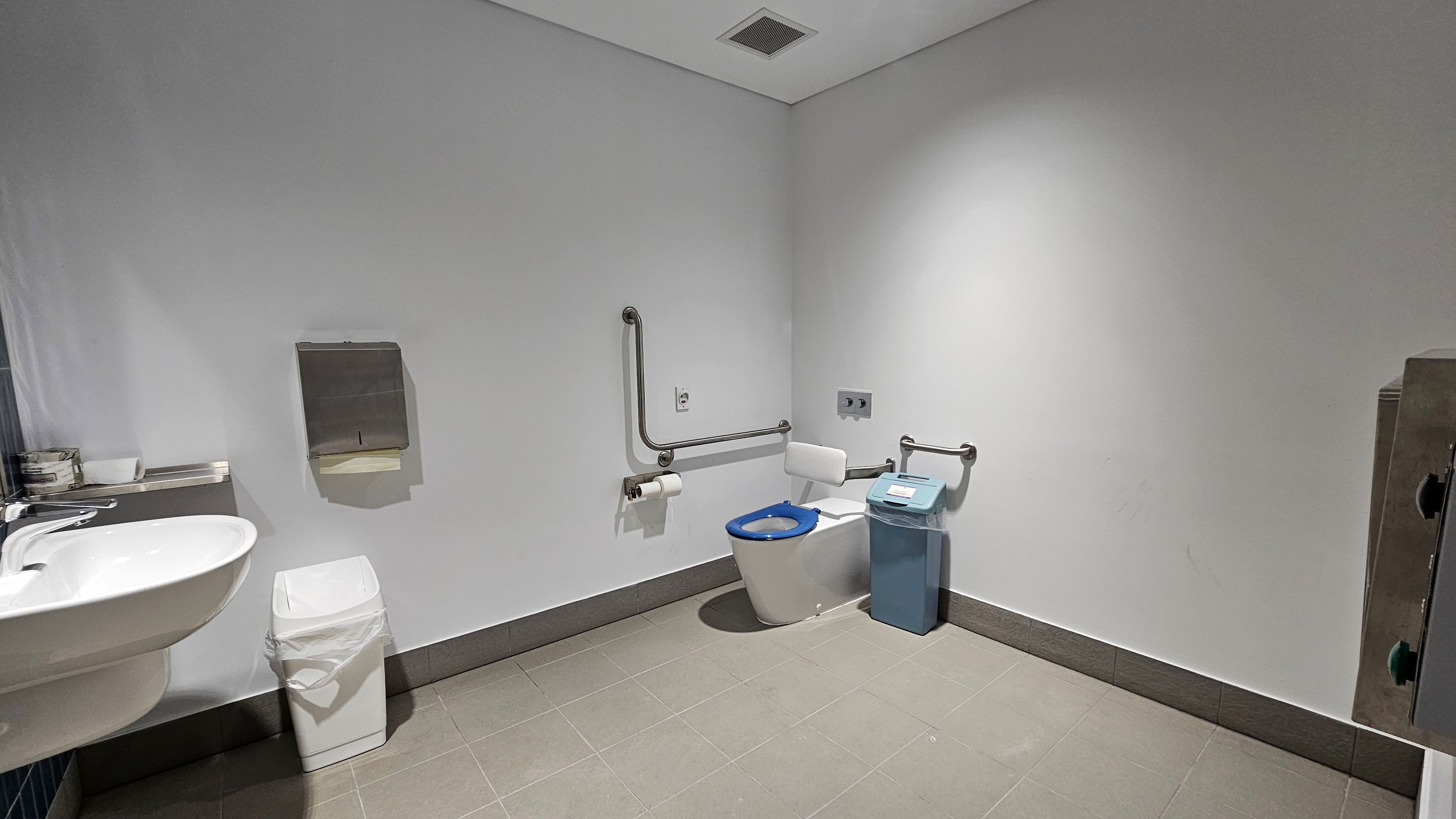 A clean, accessible bathroom with a white toilet featuring a blue seat and support bars on the right. A sanitary bin is next to the toilet. On the left, a white sink with a soap dispenser and a small bin below. A paper towel dispenser is mounted on the wall above the bin. The floor is tiled in light green, and the walls are plain white. The ceiling is white with a ventilation grille. A clean, accessible bathroom with a white toilet featuring a blue seat and support bars on the right. A sanitary bin is next to the toilet. On the left, a white sink with a soap dispenser and a small bin below. A paper towel dispenser is mounted on the wall above the bin. The floor is tiled in light green, and the walls are plain white. The ceiling is white with a ventilation grille.