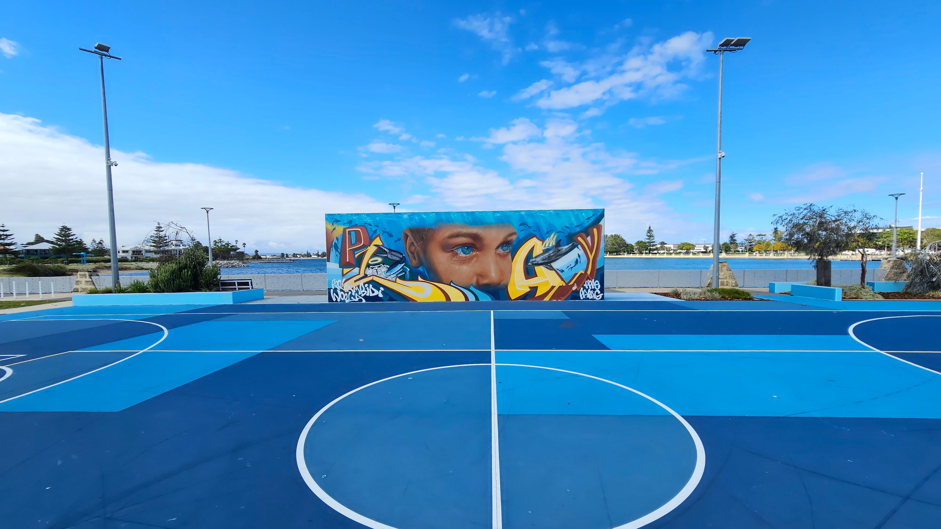 A vibrant outdoor basketball court with various shades of blue on the floor. In the centre, a large mural features a face with blue eyes, surrounded by colourful graffiti. The background shows a body of water with trees and buildings along the shore. Tall streetlights are positioned on either side of the court, and there are benches and greenery around the perimeter. A vibrant outdoor basketball court with various shades of blue on the floor. In the centre, a large mural features a face with blue eyes, surrounded by colourful graffiti. The background shows a body of water with trees and buildings along the shore. Tall streetlights are positioned on either side of the court, and there are benches and greenery around the perimeter.