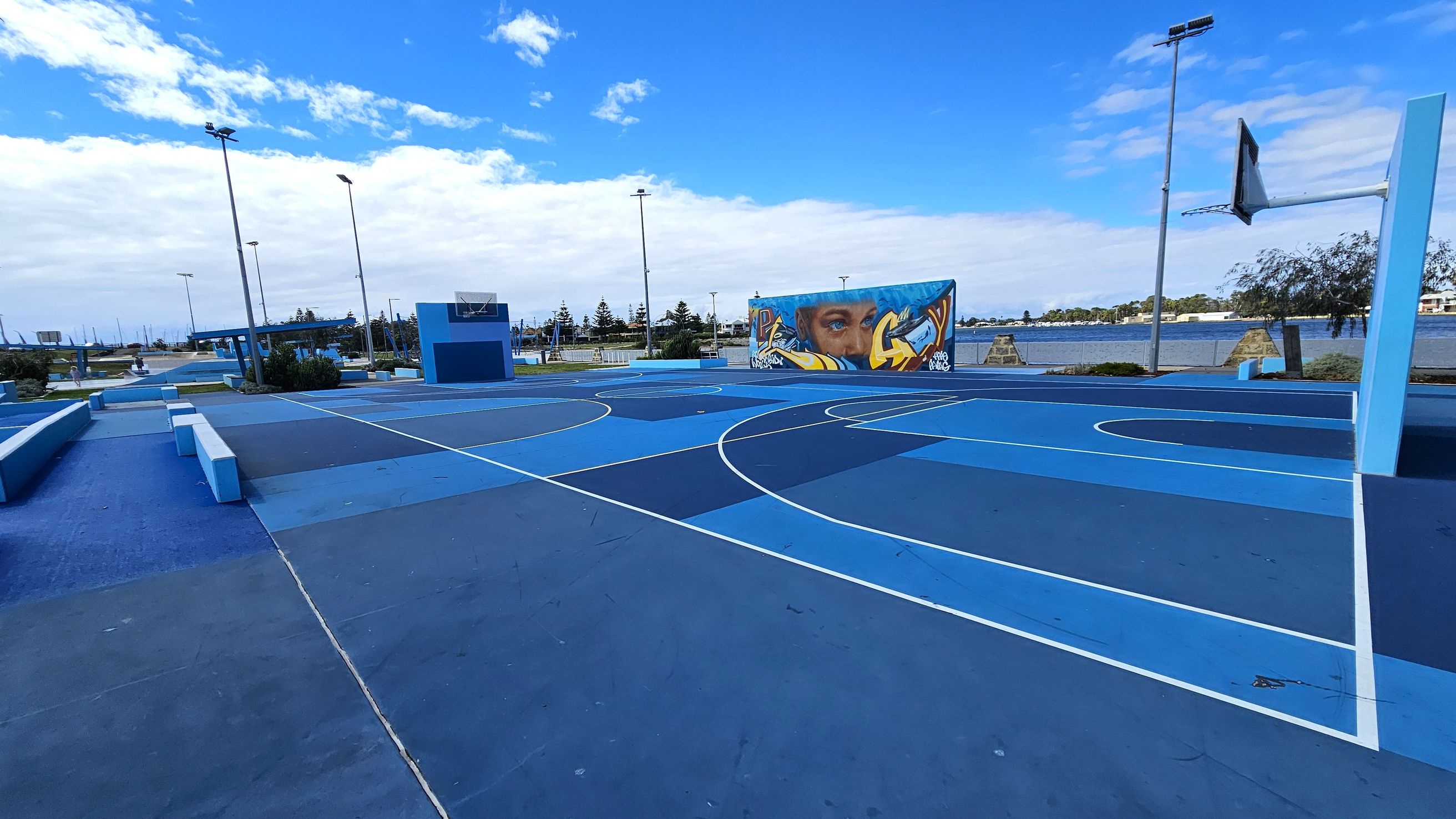 A vibrant outdoor basketball court with various shades of blue on the ground. On the right, a basketball hoop stands tall. In the background, a mural featuring a face and graffiti art is visible on a wall. Several tall streetlights are scattered around the area. Beyond the court, there is a body of water and some trees, with a few buildings in the distance. The court is surrounded by low benches and planters. A vibrant outdoor basketball court with various shades of blue on the ground. On the right, a basketball hoop stands tall. In the background, a mural featuring a face and graffiti art is visible on a wall. Several tall streetlights are scattered around the area. Beyond the court, there is a body of water and some trees, with a few buildings in the distance. The court is surrounded by low benches and planters.