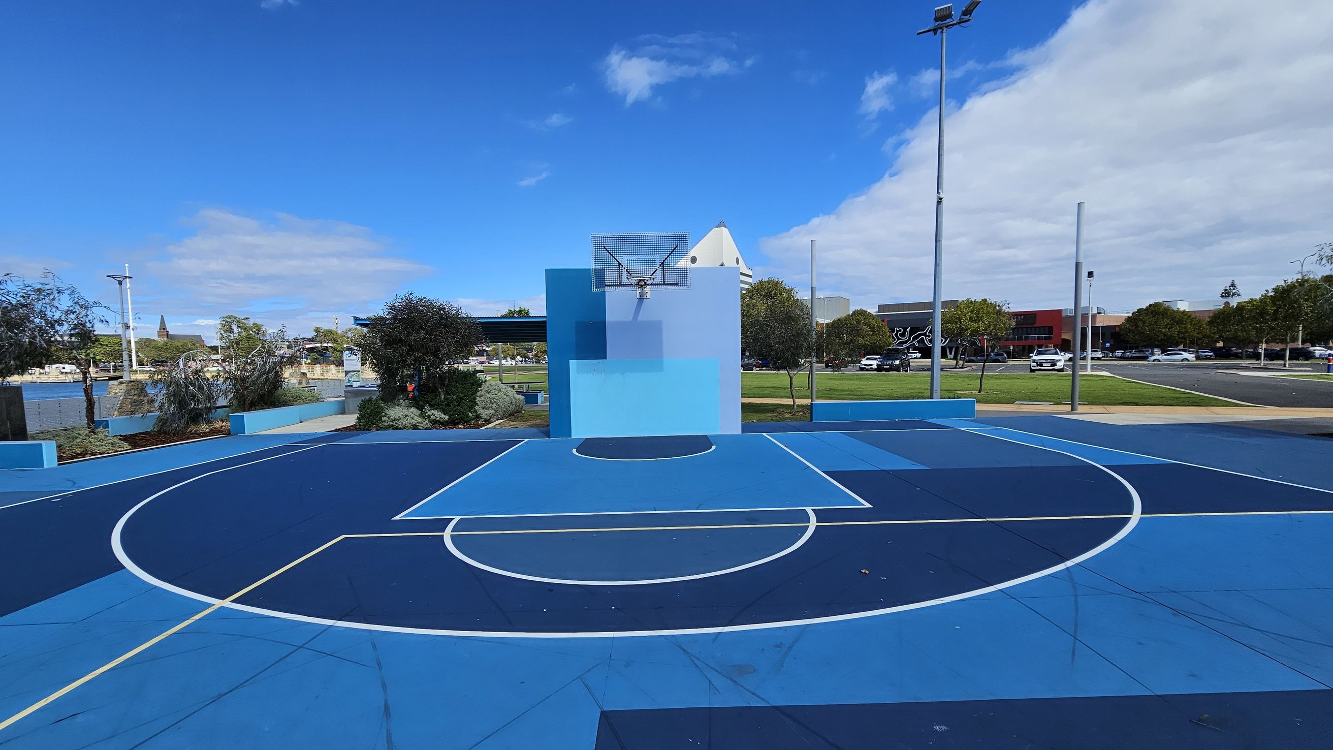 A vibrant outdoor basketball court with various shades of blue on the floor and backboard. The court features white lines marking the playing areas. In the background, there are trees and a grassy area, with a few parked cars and buildings visible. A tall streetlight stands on the right side. The sky is clear with some clouds. A vibrant outdoor basketball court with various shades of blue on the floor and backboard. The court features white lines marking the playing areas. In the background, there are trees and a grassy area, with a few parked cars and buildings visible. A tall streetlight stands on the right side. The sky is clear with some clouds.