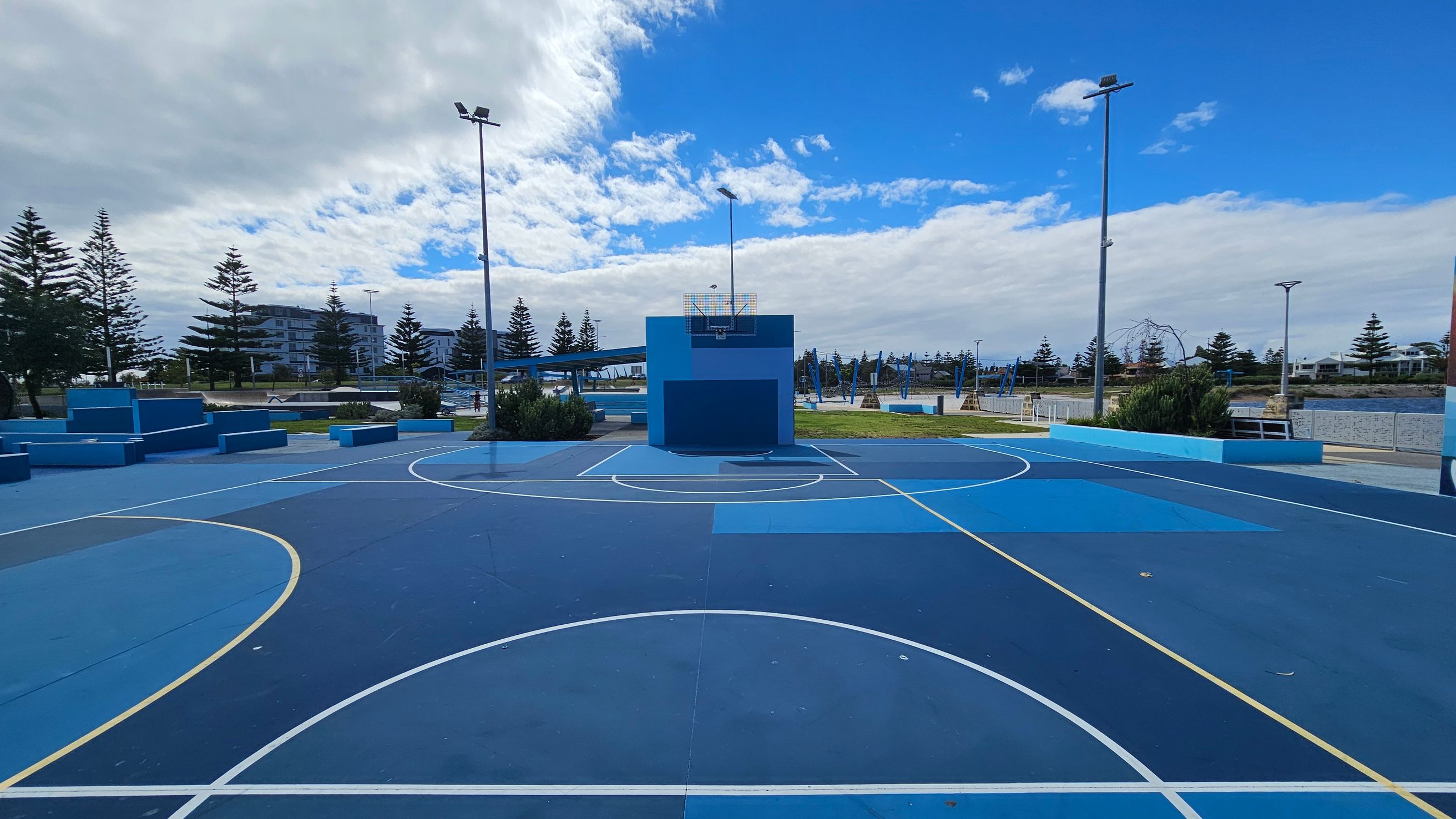 A blue outdoor basketball court with white and yellow lines. In the centre, a blue backboard and hoop are mounted on a blue wall. Surrounding the court are tall streetlights and some low bushes. In the background, there are several pine trees and modern buildings. The court surface is a mix of different shades of blue. A blue outdoor basketball court with white and yellow lines. In the centre, a blue backboard and hoop are mounted on a blue wall. Surrounding the court are tall streetlights and some low bushes. In the background, there are several pine trees and modern buildings. The court surface is a mix of different shades of blue.