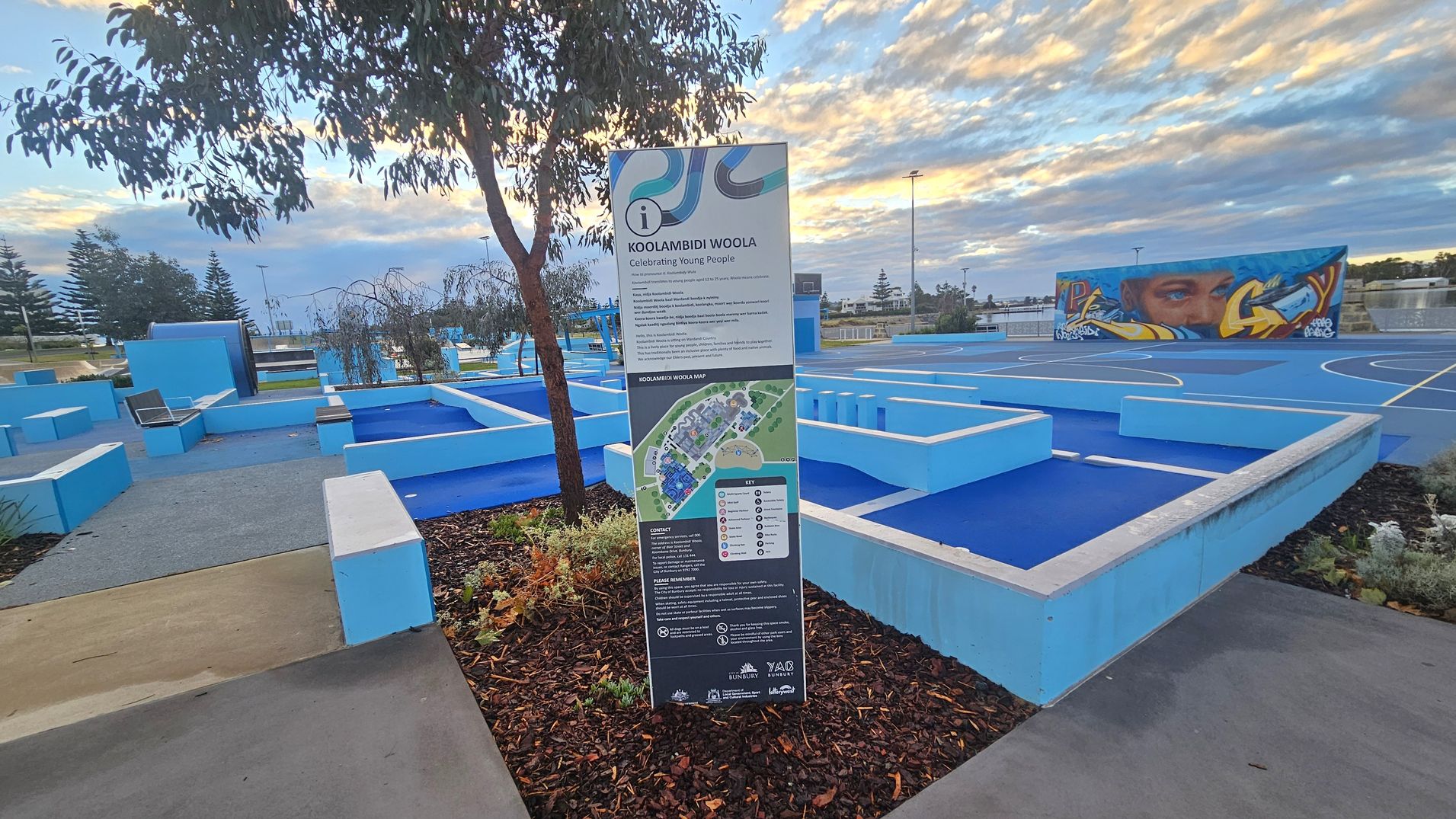 A park area with a prominent sign in the centre detailing "Koolambidi Woola" and a map. The ground is covered with blue and grey pathways and seating areas. On the right, there is a blue sports court with a mural on a wall depicting a face and abstract designs. Trees and plants are scattered throughout, and the sky is filled with clouds. A park area with a prominent sign in the centre detailing "Koolambidi Woola" and a map. The ground is covered with blue and grey pathways and seating areas. On the right, there is a blue sports court with a mural on a wall depicting a face and abstract designs. Trees and plants are scattered throughout, and the sky is filled with clouds.