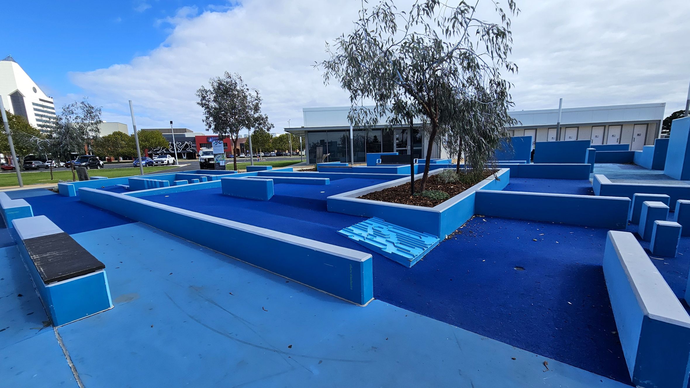 A parkour area with various blue platforms and obstacles of different heights and shapes. In the centre, there are small trees planted in raised beds. The ground is covered in blue rubberised material. In the background, there is a modern building with large windows. To the left, a street with cars and a building with a mural can be seen. The sky is partly cloudy. A parkour area with various blue platforms and obstacles of different heights and shapes. In the centre, there are small trees planted in raised beds. The ground is covered in blue rubberised material. In the background, there is a modern building with large windows. To the left, a street with cars and a building with a mural can be seen. The sky is partly cloudy.