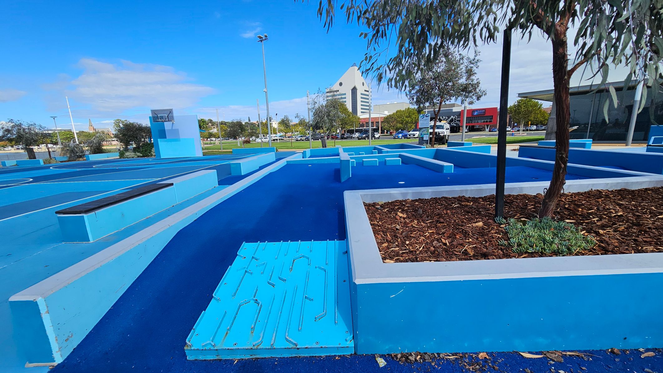 A vibrant blue outdoor area with geometric structures and pathways. On the right, a tree is planted in a raised bed filled with mulch. In the background, a distinctive triangular-roofed building is visible, along with other buildings and parked cars. The ground is covered in a textured blue surface, and there are several light poles scattered throughout the area. A vibrant blue outdoor area with geometric structures and pathways. On the right, a tree is planted in a raised bed filled with mulch. In the background, a distinctive triangular-roofed building is visible, along with other buildings and parked cars. The ground is covered in a textured blue surface, and there are several light poles scattered throughout the area.