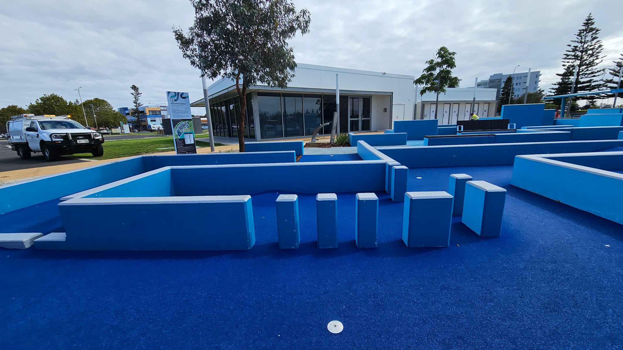 In the foreground, there is a blue outdoor play area with various geometric structures and stepping blocks. The ground is covered in blue material. In the background, a white building with large windows is visible, surrounded by a few trees. To the left, a white utility vehicle is parked on a paved area. There are signs and a grassy area near the building. Tall trees and a multi-storey building can be seen in the distance. In the foreground, there is a blue outdoor play area with various geometric structures and stepping blocks. The ground is covered in blue material. In the background, a white building with large windows is visible, surrounded by a few trees. To the left, a white utility vehicle is parked on a paved area. There are signs and a grassy area near the building. Tall trees and a multi-storey building can be seen in the distance.