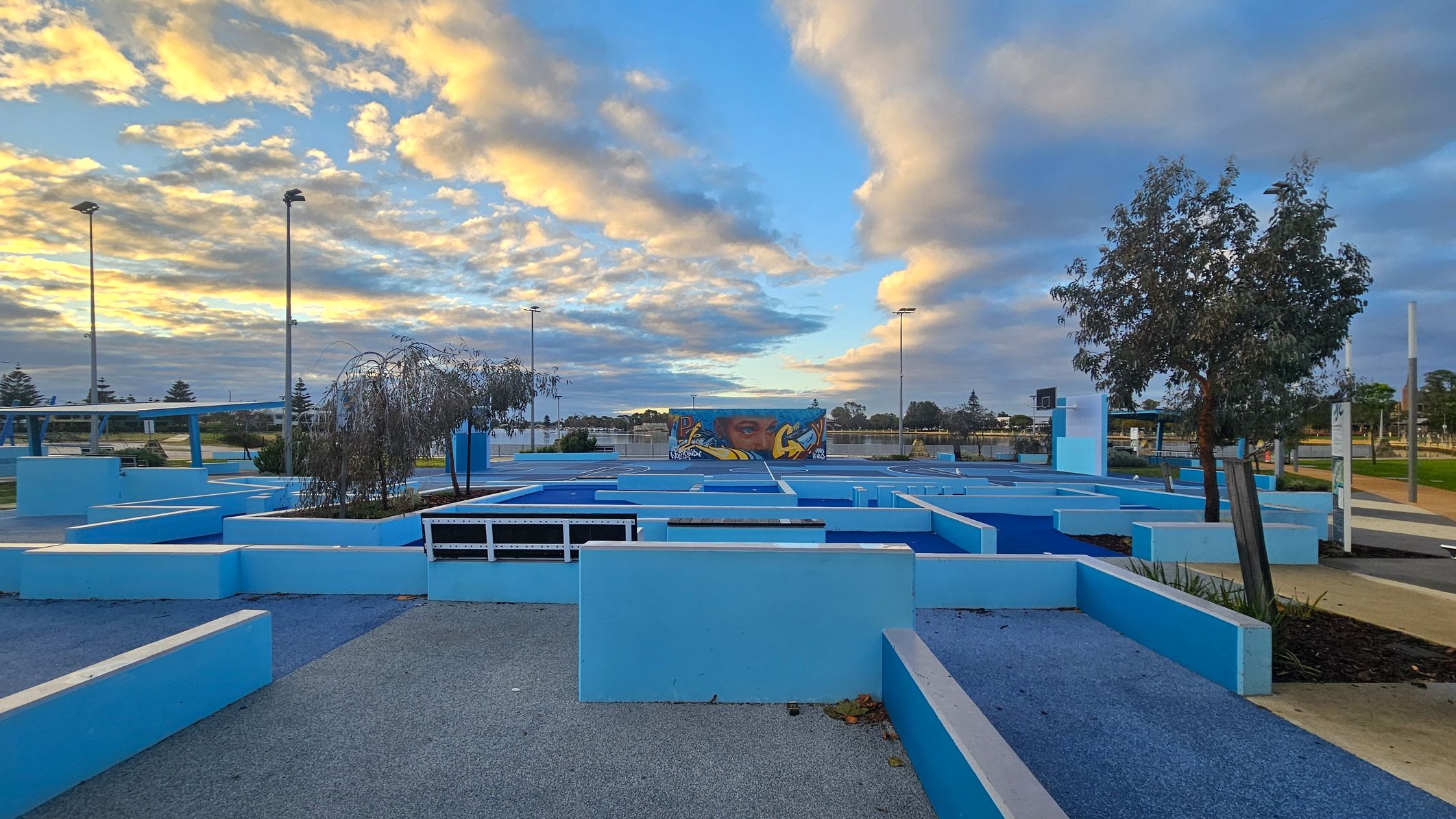A modern outdoor space with geometric blue structures and pathways. In the centre, a mural with a face and graffiti art is visible on a wall. Trees are planted within some of the structures. Tall streetlights are scattered throughout the area. The ground is a mix of grey and blue surfaces, and there are patches of greenery. The sky is filled with clouds, illuminated by the setting or rising sun. A modern outdoor space with geometric blue structures and pathways. In the centre, a mural with a face and graffiti art is visible on a wall. Trees are planted within some of the structures. Tall streetlights are scattered throughout the area. The ground is a mix of grey and blue surfaces, and there are patches of greenery. The sky is filled with clouds, illuminated by the setting or rising sun.