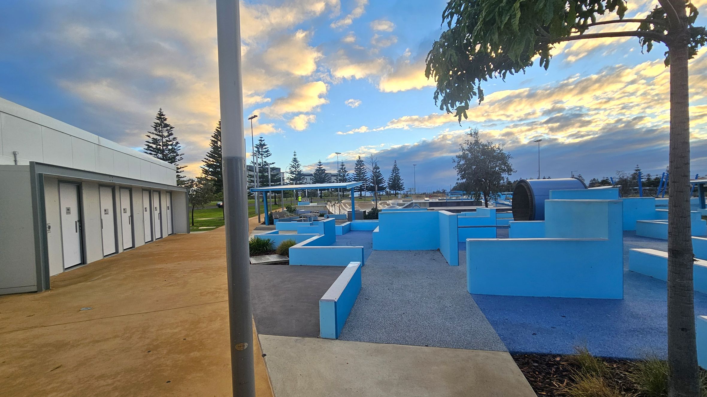 On the left, there is a row of white doors set into a grey building with a light brown concrete path in front. In the centre, a series of light blue geometric structures form a playground or park area, with a tree providing some shade. On the right, more blue structures and a large cylindrical object are visible. The background features several tall pine trees and a covered seating area. The sky is filled with clouds tinged with orange and blue hues. On the left, there is a row of white doors set into a grey building with a light brown concrete path in front. In the centre, a series of light blue geometric structures form a playground or park area, with a tree providing some shade. On the right, more blue structures and a large cylindrical object are visible. The background features several tall pine trees and a covered seating area. The sky is filled with clouds tinged with orange and blue hues.
