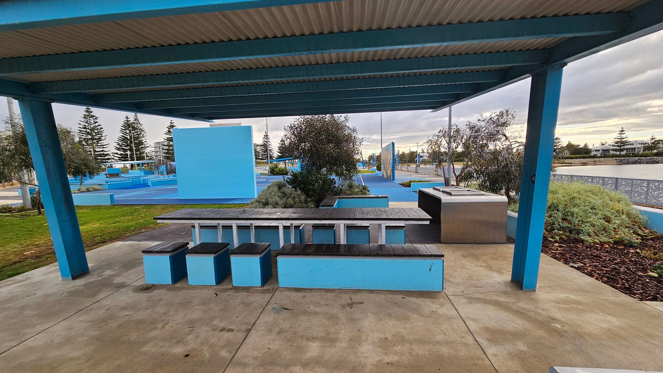 Under a blue metal shelter with a corrugated roof, there is a picnic area featuring a long table with attached benches. The benches are blue with black tops. To the right, a stainless steel barbecue is installed. The ground is a mix of concrete and grass. In the background, there are blue structures, trees, and a view of a body of water with more trees and buildings beyond. Under a blue metal shelter with a corrugated roof, there is a picnic area featuring a long table with attached benches. The benches are blue with black tops. To the right, a stainless steel barbecue is installed. The ground is a mix of concrete and grass. In the background, there are blue structures, trees, and a view of a body of water with more trees and buildings beyond.