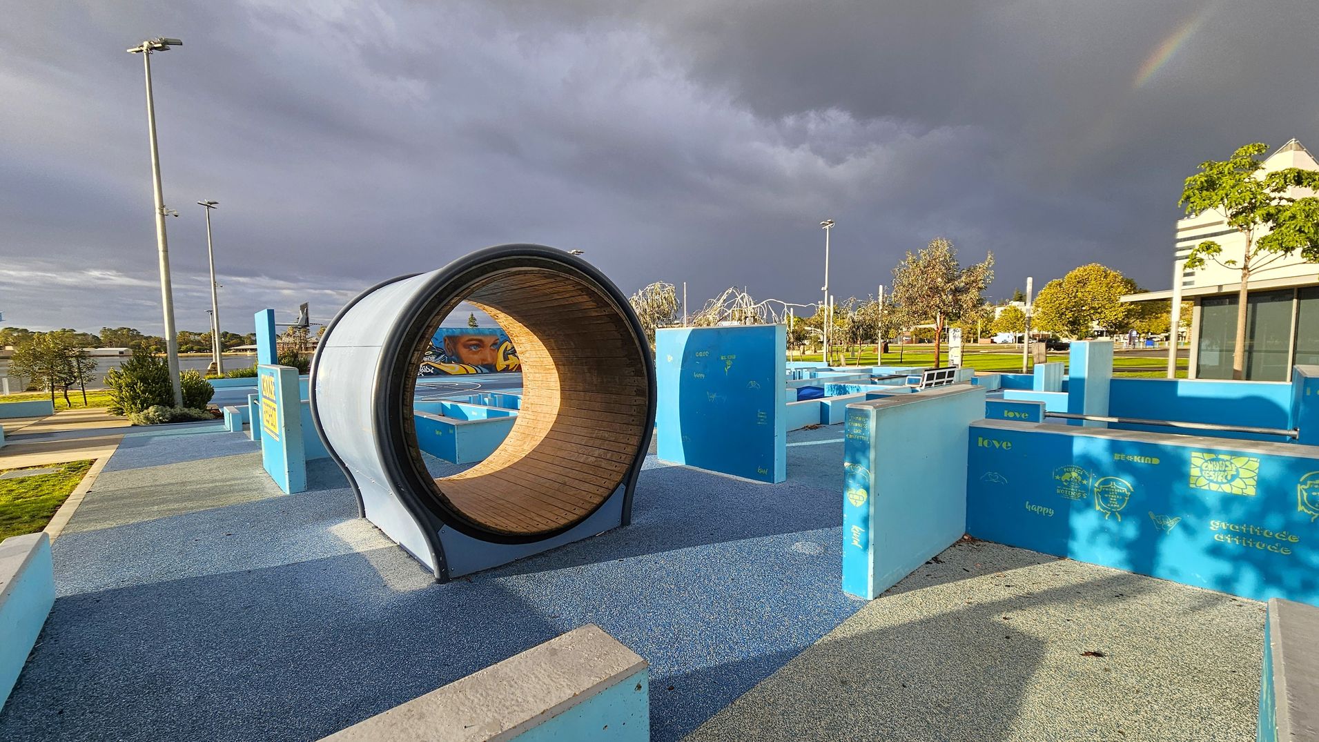 A playground with a large cylindrical tunnel in the centre, made of wood and metal. The ground is covered with blue and grey rubberised flooring. On the right, there are blue structures with words like "love" and "happy" printed on them. In the background, there are trees, a mural, and a building with a sloped roof. Tall street lamps are visible on the left, and the sky is overcast with a faint rainbow. A playground with a large cylindrical tunnel in the centre, made of wood and metal. The ground is covered with blue and grey rubberised flooring. On the right, there are blue structures with words like "love" and "happy" printed on them. In the background, there are trees, a mural, and a building with a sloped roof. Tall street lamps are visible on the left, and the sky is overcast with a faint rainbow.