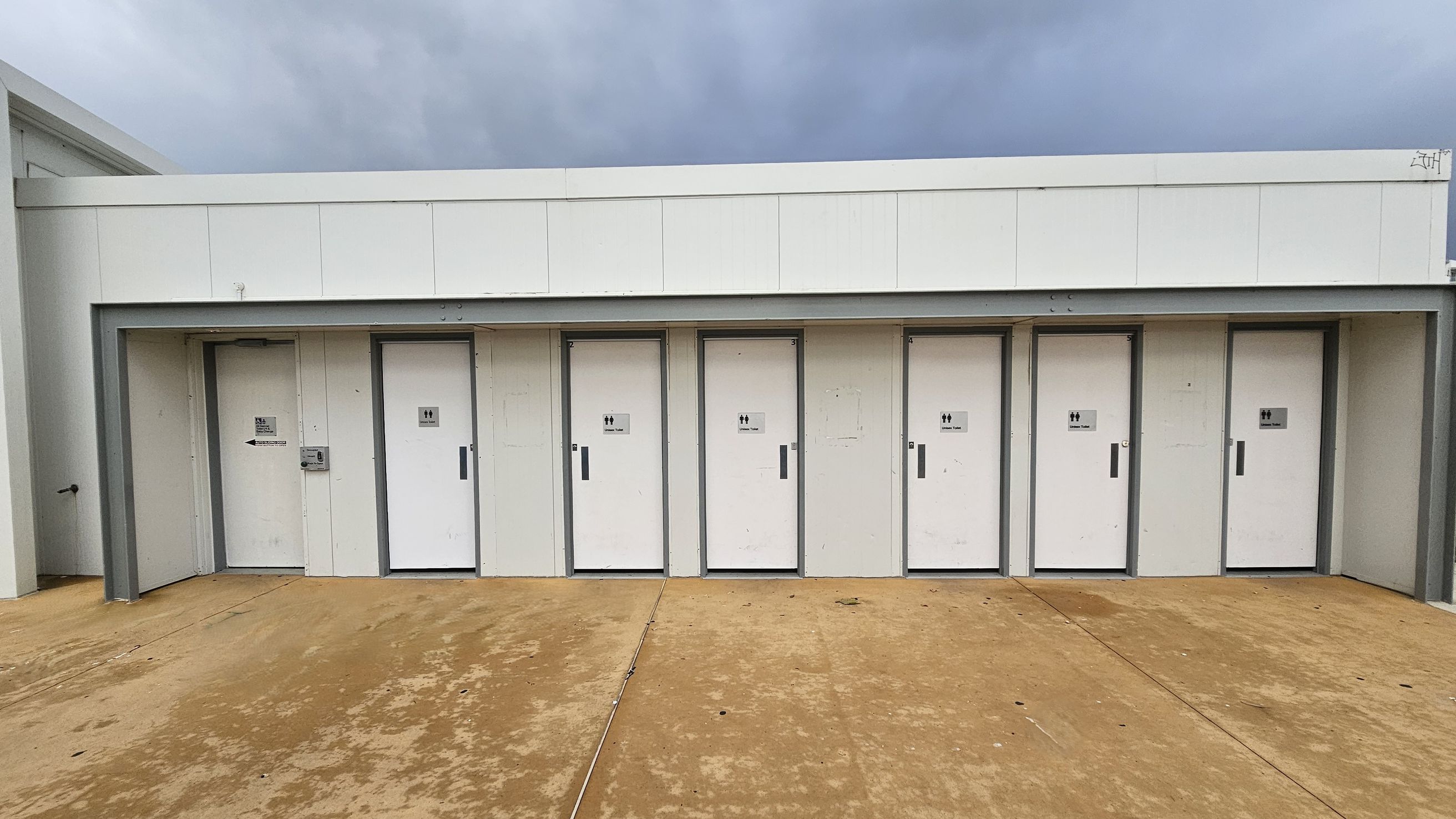 A row of six white doors, each with a unisex toilet sign, set into a white building. The doors are evenly spaced and have grey frames. The ground is a textured, light brown concrete surface. The building has a flat roof with a simple, modern design. A row of six white doors, each with a unisex toilet sign, set into a white building. The doors are evenly spaced and have grey frames. The ground is a textured, light brown concrete surface. The building has a flat roof with a simple, modern design.