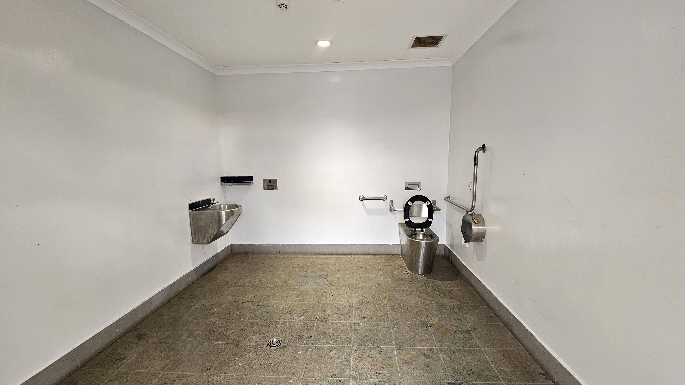 A public restroom with a stainless steel sink on the left wall, featuring a soap dispenser and a hand dryer above it. On the right, a stainless steel toilet with a black seat is installed, accompanied by grab bars for accessibility. A toilet paper dispenser is mounted nearby. The floor is tiled in brown with visible wear and stains. The ceiling is white with a recessed light and a vent. The walls are plain and painted white. A public restroom with a stainless steel sink on the left wall, featuring a soap dispenser and a hand dryer above it. On the right, a stainless steel toilet with a black seat is installed, accompanied by grab bars for accessibility. A toilet paper dispenser is mounted nearby. The floor is tiled in brown with visible wear and stains. The ceiling is white with a recessed light and a vent. The walls are plain and painted white.