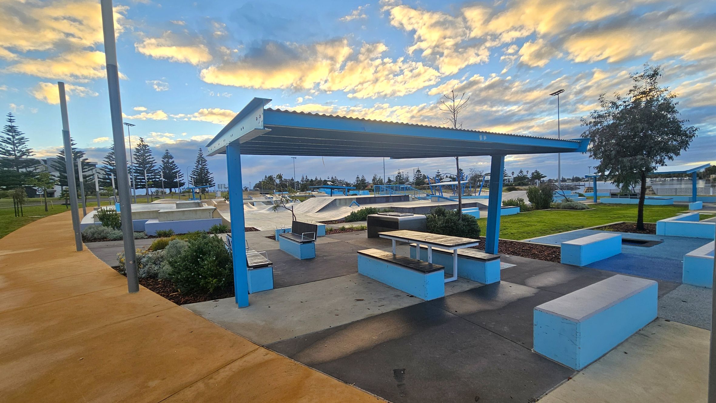 A skate park with a covered seating area in the foreground, featuring blue and white benches and tables. The ground is a mix of concrete and grass, with a yellow pathway on the left. In the background, there are skate ramps and more seating areas. Tall trees and lampposts are visible, with a line of pine trees further back. The sky is partly cloudy with a warm glow. A skate park with a covered seating area in the foreground, featuring blue and white benches and tables. The ground is a mix of concrete and grass, with a yellow pathway on the left. In the background, there are skate ramps and more seating areas. Tall trees and lampposts are visible, with a line of pine trees further back. The sky is partly cloudy with a warm glow.
