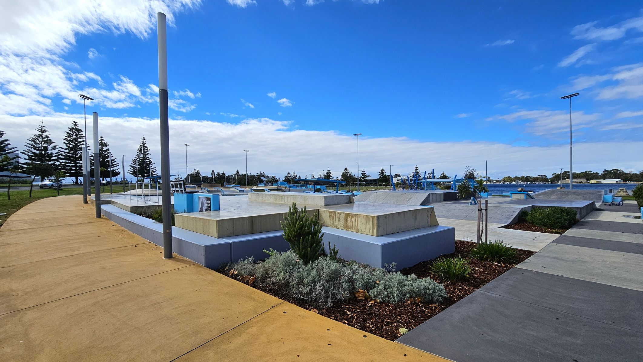 A skate park with various ramps and platforms is in the centre. On the left, a curved, tan-coloured path runs alongside tall metal poles and a row of trees. On the right, there are more ramps and a grey pathway. In the foreground, there are landscaped areas with shrubs and small plants. In the background, more trees and a clear blue sky are visible. A skate park with various ramps and platforms is in the centre. On the left, a curved, tan-coloured path runs alongside tall metal poles and a row of trees. On the right, there are more ramps and a grey pathway. In the foreground, there are landscaped areas with shrubs and small plants. In the background, more trees and a clear blue sky are visible.