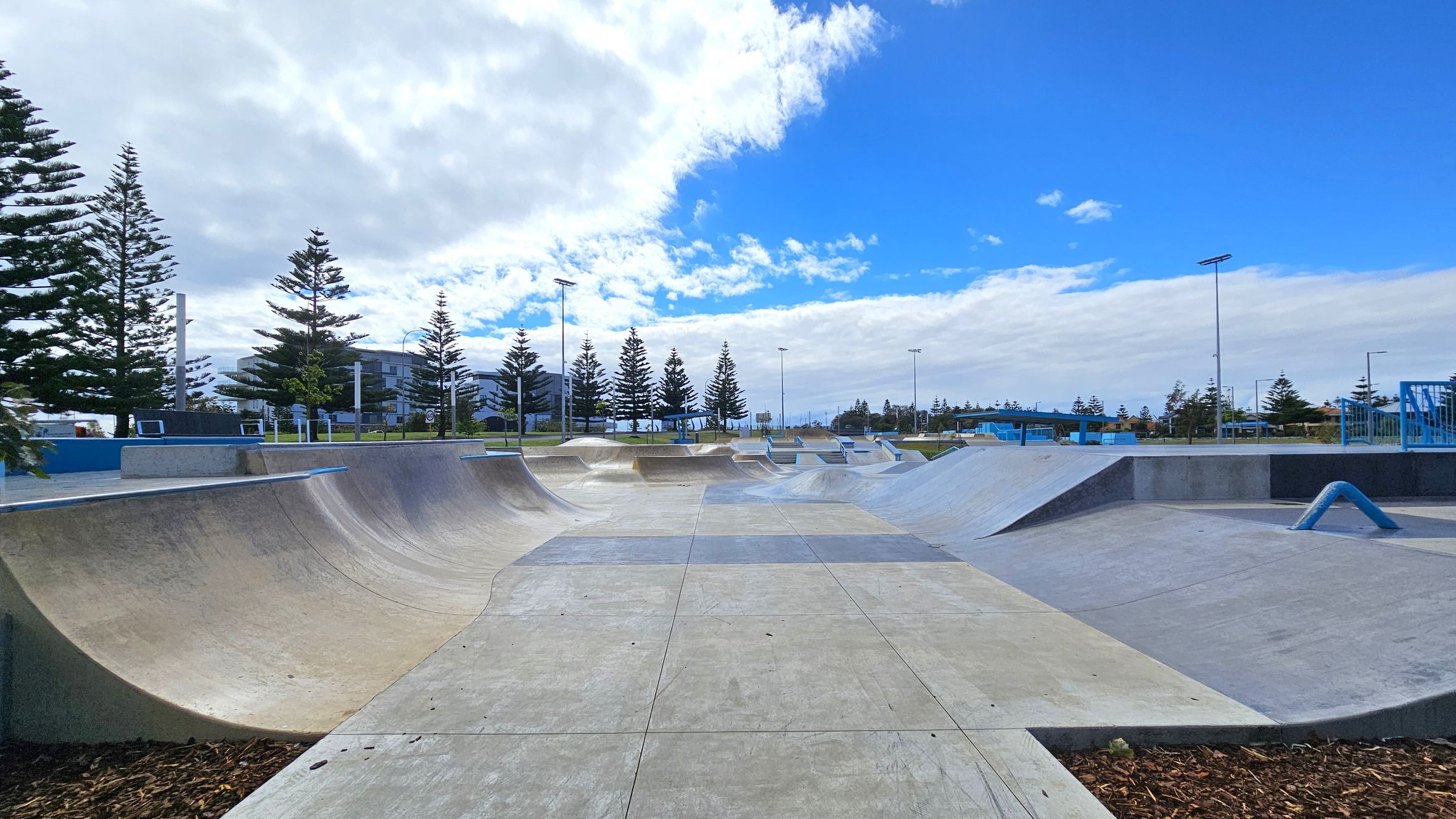 A concrete skate park with various ramps and curves is in the foreground. On the left, there are tall pine trees and a building in the background. On the right, more ramps and a blue rail are visible. The park is surrounded by more trees and light poles. The sky is partly cloudy with patches of blue. A concrete skate park with various ramps and curves is in the foreground. On the left, there are tall pine trees and a building in the background. On the right, more ramps and a blue rail are visible. The park is surrounded by more trees and light poles. The sky is partly cloudy with patches of blue.