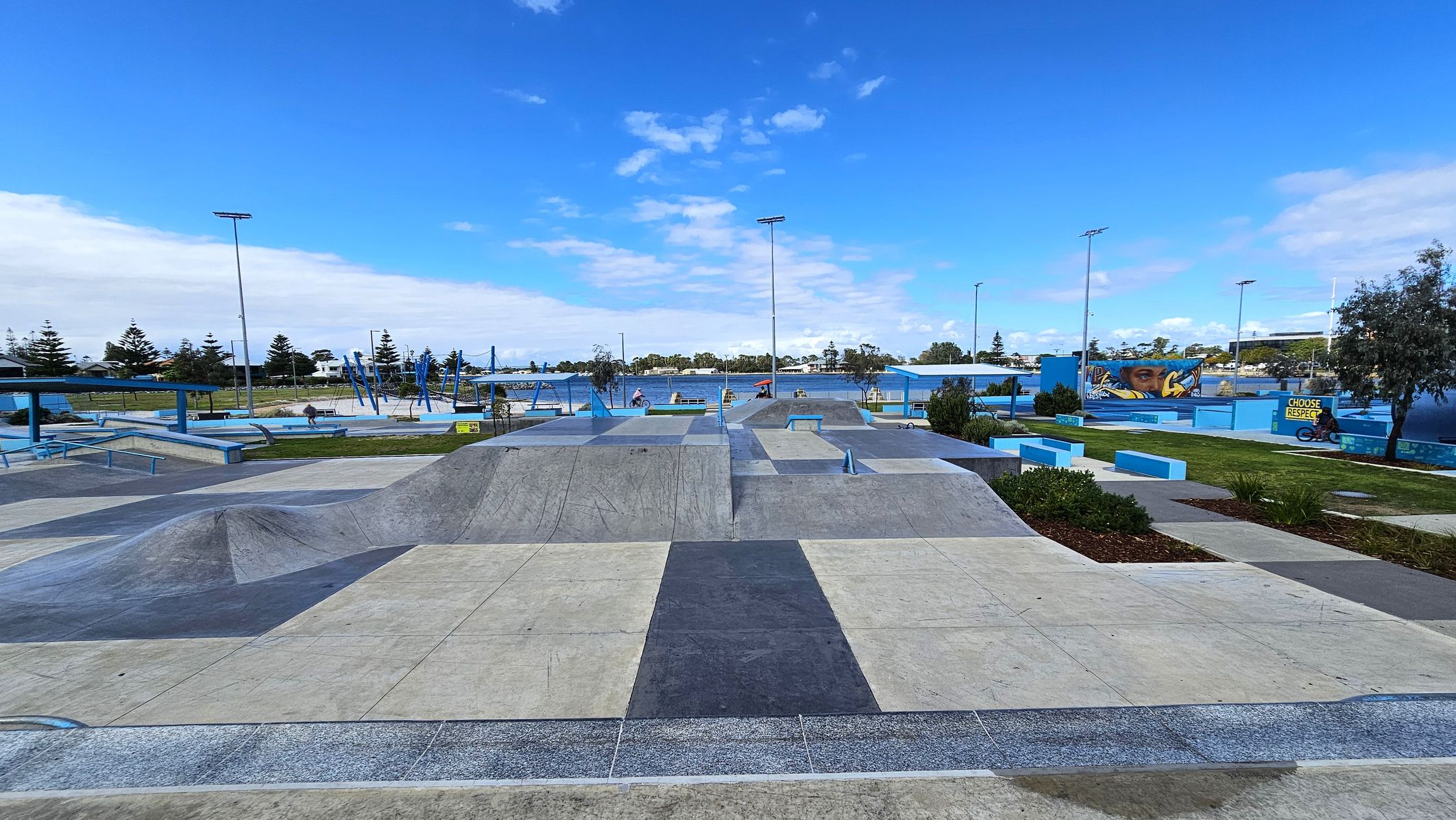 A skate park with various ramps and rails in the foreground, featuring a mix of concrete and blue-painted surfaces. In the background, there are trees and a mural with a face on a wall. To the right, a person on a bicycle is near a sign that reads "CHOOSE RESPECT". The area is open and spacious, with scattered greenery and a clear sky above. A skate park with various ramps and rails in the foreground, featuring a mix of concrete and blue-painted surfaces. In the background, there are trees and a mural with a face on a wall. To the right, a person on a bicycle is near a sign that reads "CHOOSE RESPECT". The area is open and spacious, with scattered greenery and a clear sky above.