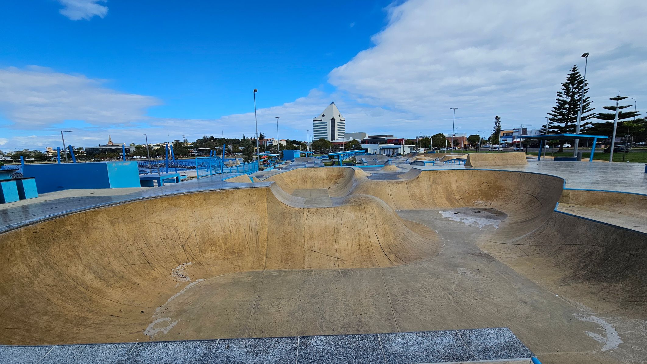 A skate park with large concrete bowls and ramps in the foreground. The surface is worn with visible marks. In the background, there is a distinctive white building with a triangular top. To the right, tall pine trees and street lamps line the area. Blue railings and benches are scattered throughout the park. The sky is partly cloudy, and the surrounding area includes various buildings and greenery. A skate park with large concrete bowls and ramps in the foreground. The surface is worn with visible marks. In the background, there is a distinctive white building with a triangular top. To the right, tall pine trees and street lamps line the area. Blue railings and benches are scattered throughout the park. The sky is partly cloudy, and the surrounding area includes various buildings and greenery.