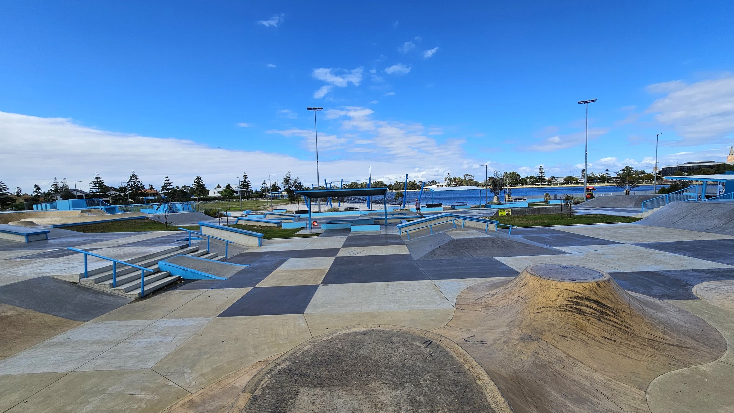 A skate park with various ramps, rails, and platforms in shades of blue and grey. The ground is a patchwork of different coloured concrete sections. In the background, there are trees and a body of water. Several tall light poles are visible, and a few people are scattered throughout the park. The area is open and spacious, with a clear view of the surroundings. A skate park with various ramps, rails, and platforms in shades of blue and grey. The ground is a patchwork of different coloured concrete sections. In the background, there are trees and a body of water. Several tall light poles are visible, and a few people are scattered throughout the park. The area is open and spacious, with a clear view of the surroundings.