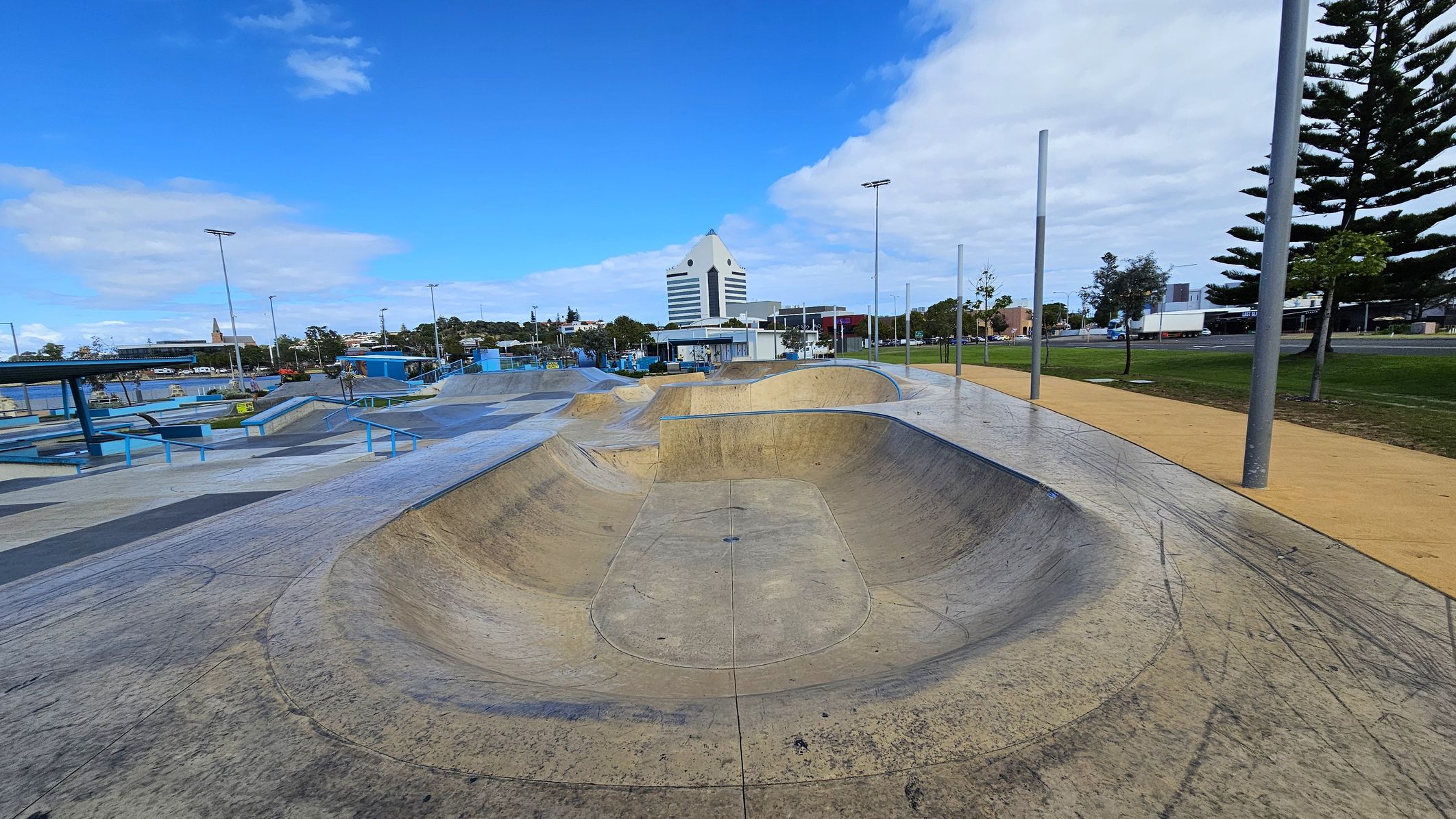 A skate park with a large concrete bowl in the foreground, featuring various ramps and rails. On the left, there are blue structures and benches. In the background, a distinctive white building with a triangular top is visible, surrounded by trees and other buildings. On the right, there is a grassy area with tall trees and a path lined with streetlights. The sky is mostly clear with some clouds. A skate park with a large concrete bowl in the foreground, featuring various ramps and rails. On the left, there are blue structures and benches. In the background, a distinctive white building with a triangular top is visible, surrounded by trees and other buildings. On the right, there is a grassy area with tall trees and a path lined with streetlights. The sky is mostly clear with some clouds.