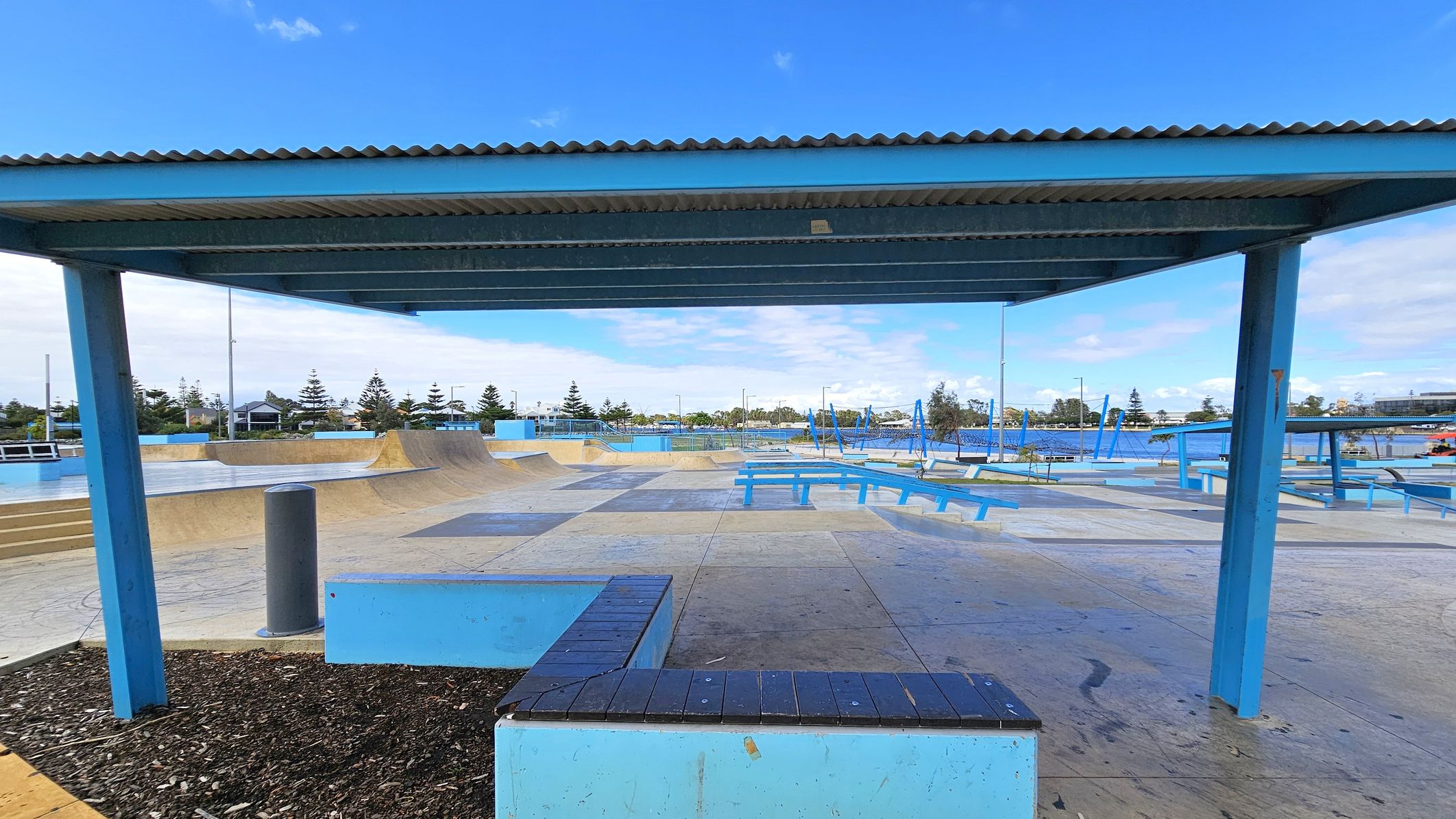A skate park with various ramps and benches is visible. In the foreground, there's a blue metal shelter with a corrugated roof supported by blue pillars. The ground is a mix of concrete and wooden surfaces. In the background, there are trees and some buildings. Blue benches and additional structures are scattered throughout the park. The area is open and spacious, designed for skateboarding activities. A skate park with various ramps and benches is visible. In the foreground, there's a blue metal shelter with a corrugated roof supported by blue pillars. The ground is a mix of concrete and wooden surfaces. In the background, there are trees and some buildings. Blue benches and additional structures are scattered throughout the park. The area is open and spacious, designed for skateboarding activities.
