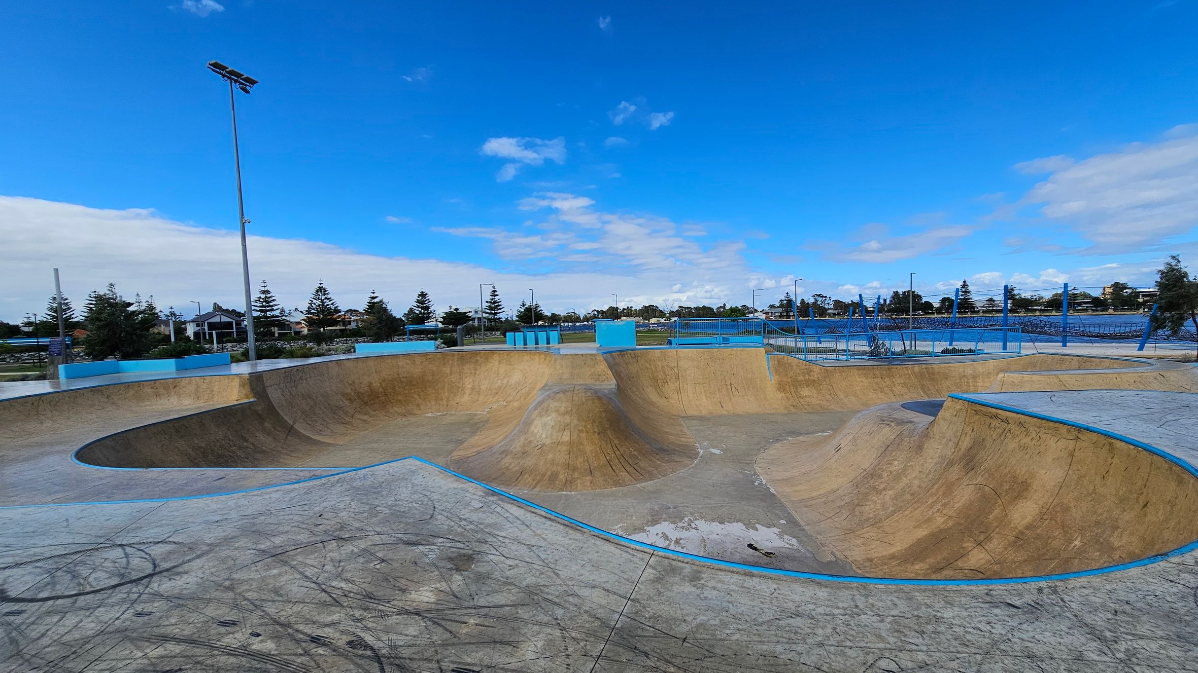 A skate park with multiple ramps and bowls in the foreground, featuring a smooth concrete surface with visible marks and scratches. The ramps are bordered with blue edges. In the background, there are tall floodlights and a fence enclosing the area. Beyond the fence, trees and buildings are visible, with a clear blue sky above. A skate park with multiple ramps and bowls in the foreground, featuring a smooth concrete surface with visible marks and scratches. The ramps are bordered with blue edges. In the background, there are tall floodlights and a fence enclosing the area. Beyond the fence, trees and buildings are visible, with a clear blue sky above.