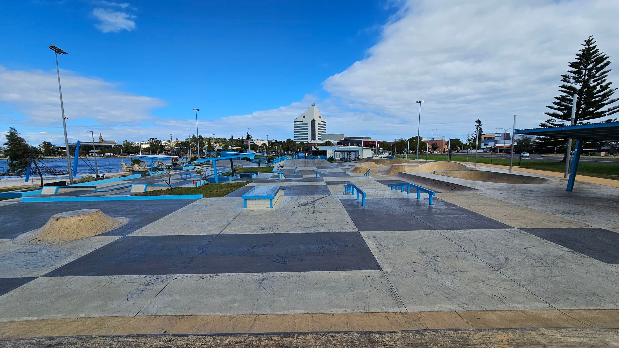 A skate park with various ramps, rails, and platforms in the foreground. The ground is a patchwork of different coloured concrete sections. In the background, there is a large, distinctive white building with a triangular top. To the left, there is a body of water with trees and a tall structure visible. On the right, there are more trees and a few buildings. The sky is partly cloudy, and there are tall streetlights throughout the area. A skate park with various ramps, rails, and platforms in the foreground. The ground is a patchwork of different coloured concrete sections. In the background, there is a large, distinctive white building with a triangular top. To the left, there is a body of water with trees and a tall structure visible. On the right, there are more trees and a few buildings. The sky is partly cloudy, and there are tall streetlights throughout the area.