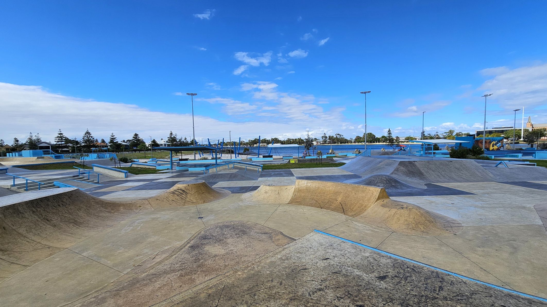 A skate park with various ramps and rails is in the foreground, featuring a mix of concrete and metal structures. The middle ground has more skate features and a few trees. In the background, there are tall light poles, a body of water, and a row of trees. A mural is visible on the right side. The sky is clear with some clouds. A skate park with various ramps and rails is in the foreground, featuring a mix of concrete and metal structures. The middle ground has more skate features and a few trees. In the background, there are tall light poles, a body of water, and a row of trees. A mural is visible on the right side. The sky is clear with some clouds.