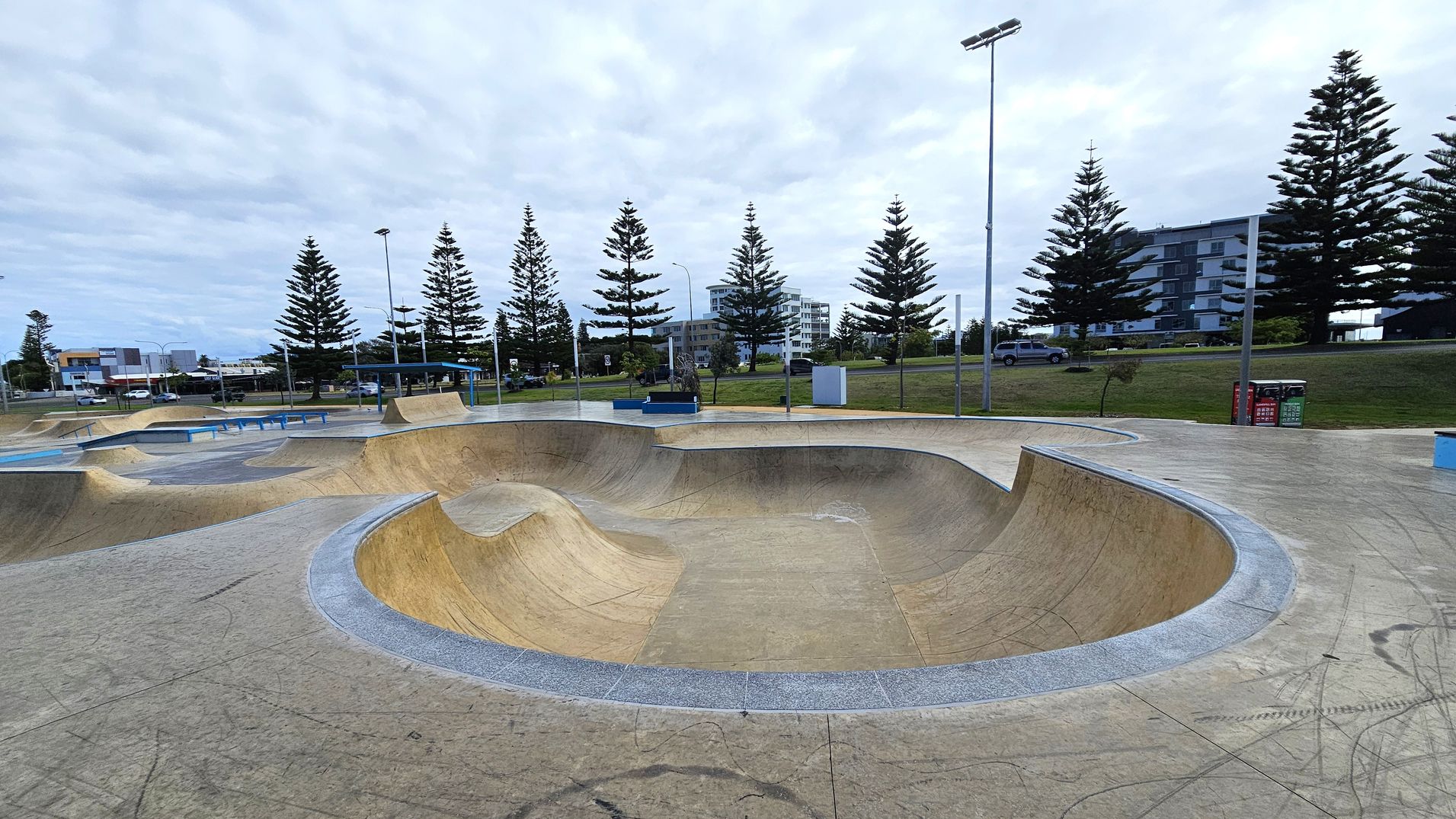 A skate park with smooth concrete surfaces and curved ramps in the foreground. In the background, there are tall pine trees lining the area, with a few modern buildings visible beyond them. The ground is marked with various lines and scuffs from skateboards. There are street lamps and a few benches scattered around the park. A couple of bins are positioned on the right side near the trees. A skate park with smooth concrete surfaces and curved ramps in the foreground. In the background, there are tall pine trees lining the area, with a few modern buildings visible beyond them. The ground is marked with various lines and scuffs from skateboards. There are street lamps and a few benches scattered around the park. A couple of bins are positioned on the right side near the trees.