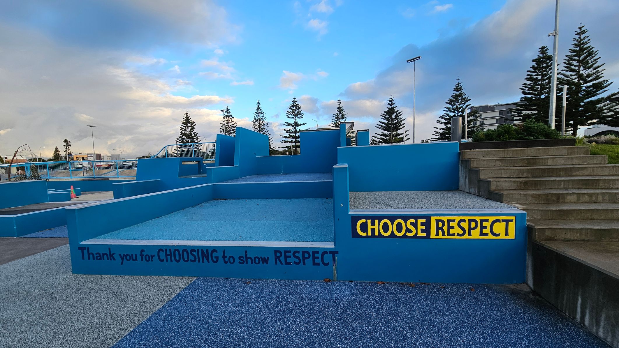 A blue concrete structure with multiple levels and steps is in the centre. On the right, a set of grey steps leads up to a grassy area. A yellow and black sign reads "CHOOSE RESPECT." Another message on the structure says, "Thank you for CHOOSING to show RESPECT." In the background, there are tall pine trees and a few buildings. The ground is covered with a textured blue surface, and there are railings and a traffic cone visible on the left. A blue concrete structure with multiple levels and steps is in the centre. On the right, a set of grey steps leads up to a grassy area. A yellow and black sign reads "CHOOSE RESPECT." Another message on the structure says, "Thank you for CHOOSING to show RESPECT." In the background, there are tall pine trees and a few buildings. The ground is covered with a textured blue surface, and there are railings and a traffic cone visible on the left.
