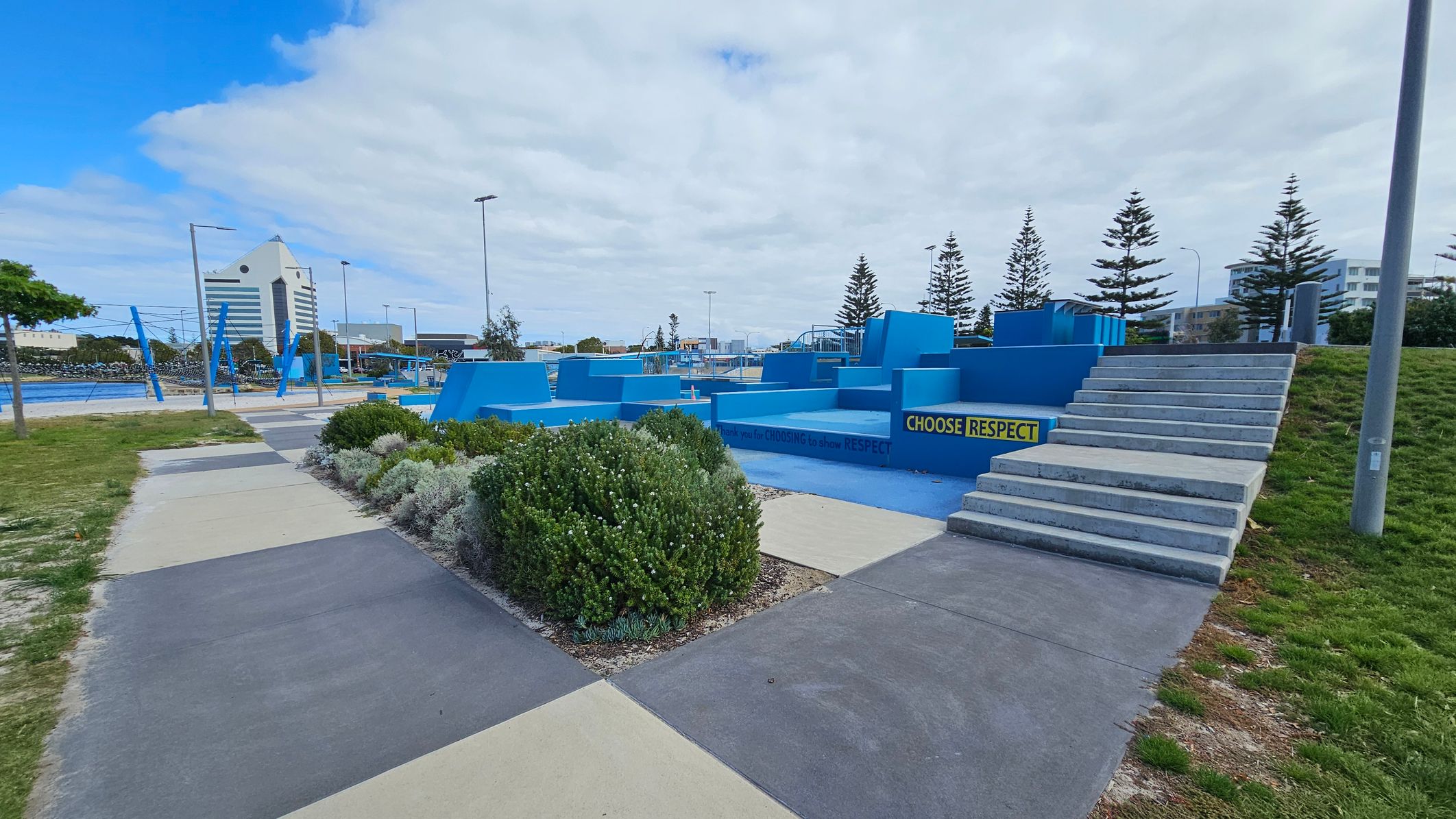 A skate park with blue ramps and structures is in the centre, featuring a sign that reads "CHOOSE RESPECT". On the right, there are concrete steps leading up to the park. In the foreground, there are patches of grass and a paved walkway with different coloured sections. In the background, a tall building with a triangular roof is visible, along with several pine trees and streetlights. A skate park with blue ramps and structures is in the centre, featuring a sign that reads "CHOOSE RESPECT". On the right, there are concrete steps leading up to the park. In the foreground, there are patches of grass and a paved walkway with different coloured sections. In the background, a tall building with a triangular roof is visible, along with several pine trees and streetlights.