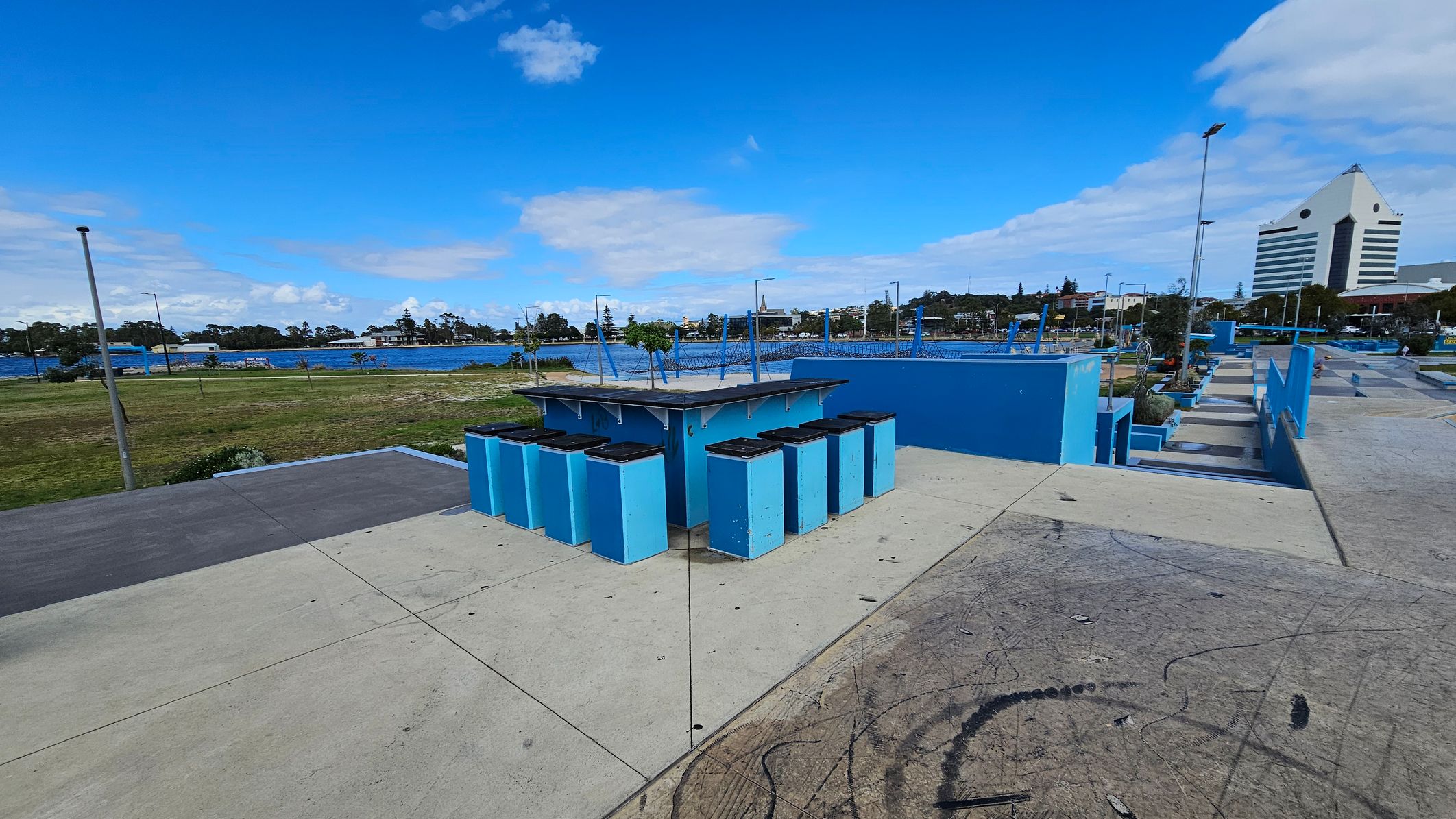 A concrete skate park with blue ramps and structures. In the foreground, there are several blue bins with black lids arranged in a semi-circle. The background features a grassy area leading to a body of water, with trees and buildings visible across the water. On the right, a distinctive white building with a triangular top stands out. The sky is clear with some clouds. A concrete skate park with blue ramps and structures. In the foreground, there are several blue bins with black lids arranged in a semi-circle. The background features a grassy area leading to a body of water, with trees and buildings visible across the water. On the right, a distinctive white building with a triangular top stands out. The sky is clear with some clouds.