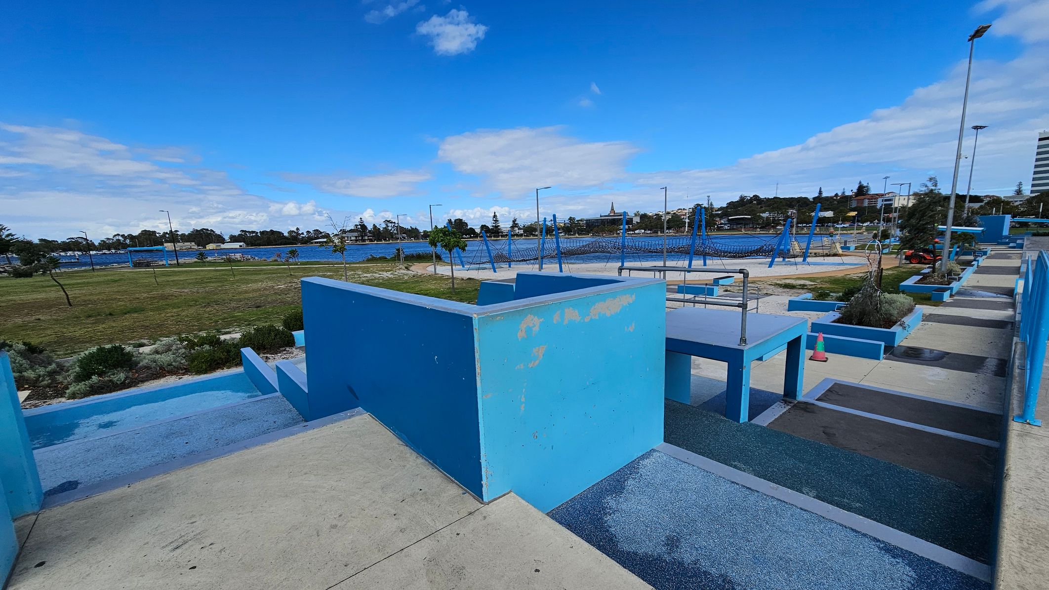 Concrete pathways and blue-painted structures are in the foreground, with a series of ramps and steps. In the middle, there are blue climbing frames and swings in a playground area. The background features a grassy field with scattered trees, a body of water, and a distant view of buildings and trees. The sky is mostly clear with some clouds. Concrete pathways and blue-painted structures are in the foreground, with a series of ramps and steps. In the middle, there are blue climbing frames and swings in a playground area. The background features a grassy field with scattered trees, a body of water, and a distant view of buildings and trees. The sky is mostly clear with some clouds.