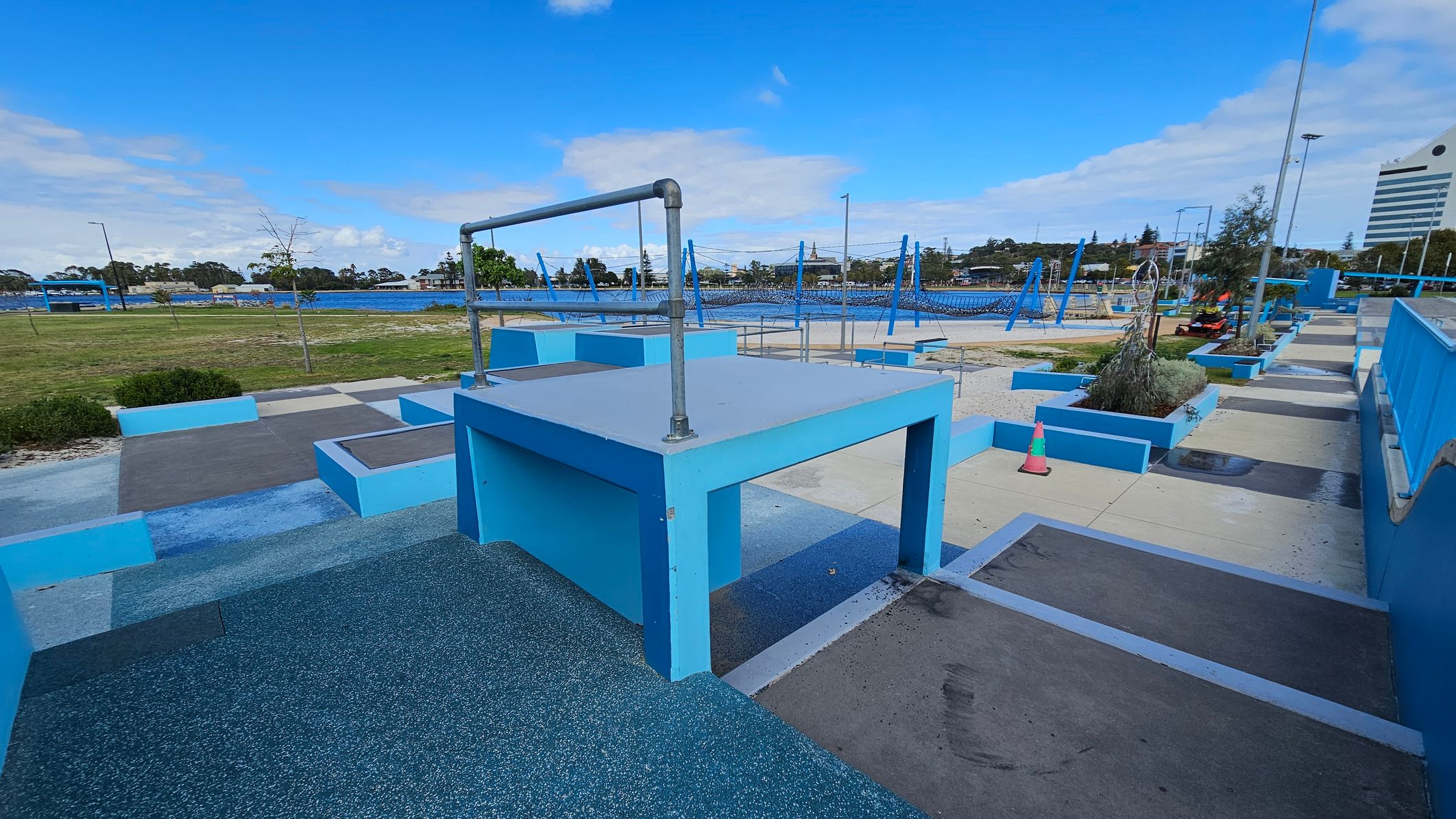 A parkour training area with various blue structures and platforms. In the foreground, a large blue platform with a metal bar on top is visible. To the right, there are more platforms and a pathway with a traffic cone. In the background, a grassy area and a body of water can be seen, along with trees and a few buildings. The ground is a mix of textured surfaces and concrete, and the area is open and spacious. A parkour training area with various blue structures and platforms. In the foreground, a large blue platform with a metal bar on top is visible. To the right, there are more platforms and a pathway with a traffic cone. In the background, a grassy area and a body of water can be seen, along with trees and a few buildings. The ground is a mix of textured surfaces and concrete, and the area is open and spacious.