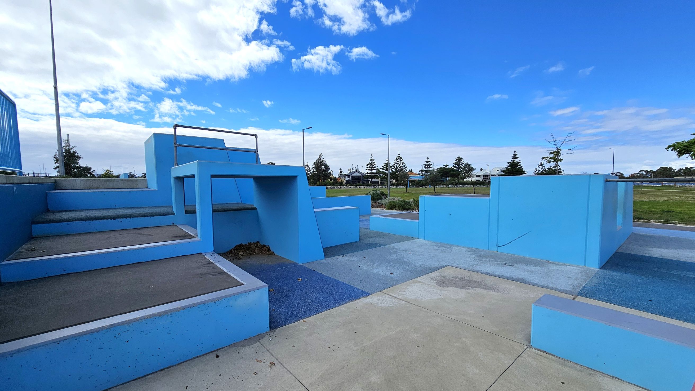 A parkour training area with various blue structures, including steps and platforms, is in the foreground. The ground is a mix of concrete and textured surfaces. In the background, there are trees, a grassy area, and a few buildings. Street lamps line the path, and the sky is partly cloudy. A parkour training area with various blue structures, including steps and platforms, is in the foreground. The ground is a mix of concrete and textured surfaces. In the background, there are trees, a grassy area, and a few buildings. Street lamps line the path, and the sky is partly cloudy.
