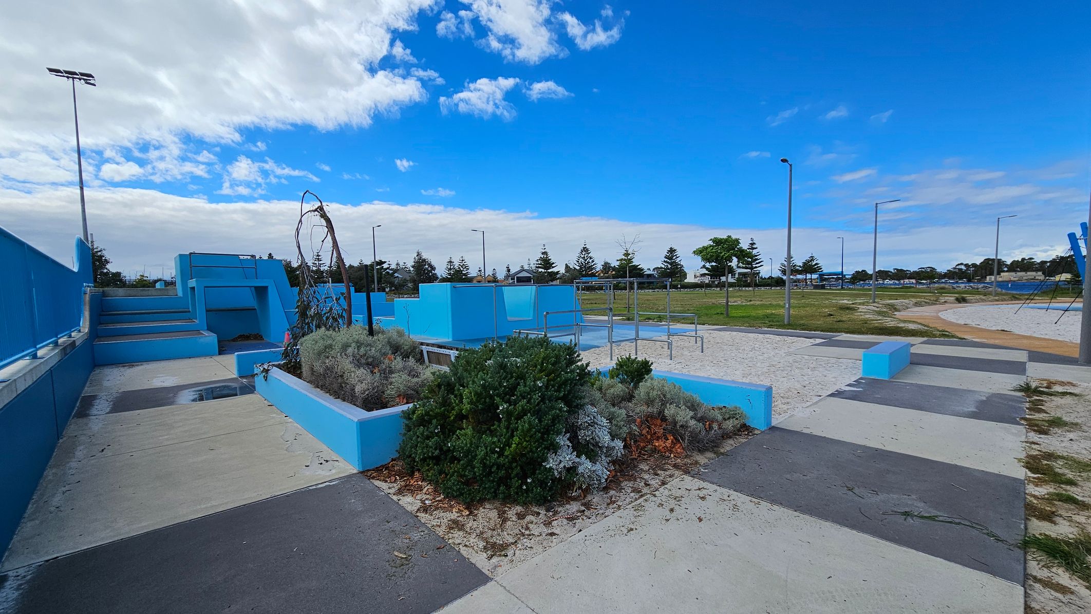 A park area with a blue and grey colour scheme. On the left, there are blue steps leading up to a platform with railings. In the centre, a raised garden bed with various shrubs and a small tree is surrounded by blue concrete borders. To the right, there is a sandy area with metal bars, possibly for exercise or play. The ground is a mix of concrete and sand, and there are several streetlights and trees in the background. A park area with a blue and grey colour scheme. On the left, there are blue steps leading up to a platform with railings. In the centre, a raised garden bed with various shrubs and a small tree is surrounded by blue concrete borders. To the right, there is a sandy area with metal bars, possibly for exercise or play. The ground is a mix of concrete and sand, and there are several streetlights and trees in the background.
