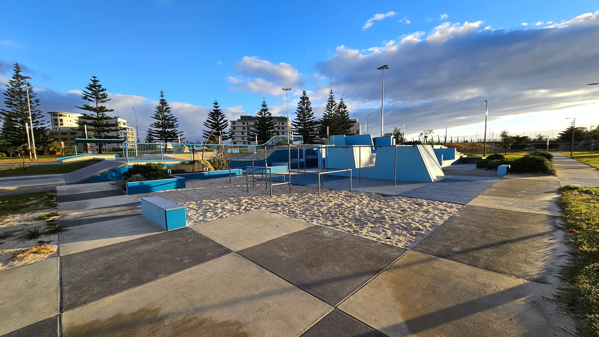 A skate park with various ramps and structures painted in blue and grey. The ground is a mix of concrete and sand. In the background, there are tall pine trees and several multi-storey buildings. The area is well-lit with tall street lamps, and there are pathways and grassy areas surrounding the park. A skate park with various ramps and structures painted in blue and grey. The ground is a mix of concrete and sand. In the background, there are tall pine trees and several multi-storey buildings. The area is well-lit with tall street lamps, and there are pathways and grassy areas surrounding the park.