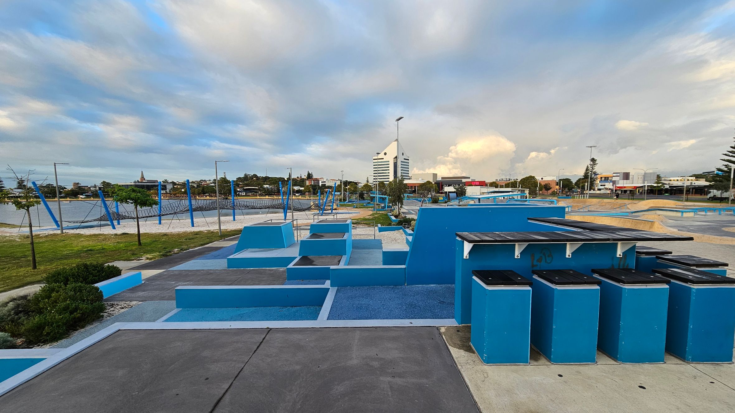 A skate park with blue ramps and structures is in the foreground, alongside a row of blue bins with black lids. In the background, there's a large building with a distinctive triangular roof. To the left, a sandy area with a netted climbing structure is visible, bordered by grass and small trees. The area is surrounded by various buildings and streetlights. The ground is a mix of concrete and blue surfaces. A skate park with blue ramps and structures is in the foreground, alongside a row of blue bins with black lids. In the background, there's a large building with a distinctive triangular roof. To the left, a sandy area with a netted climbing structure is visible, bordered by grass and small trees. The area is surrounded by various buildings and streetlights. The ground is a mix of concrete and blue surfaces.