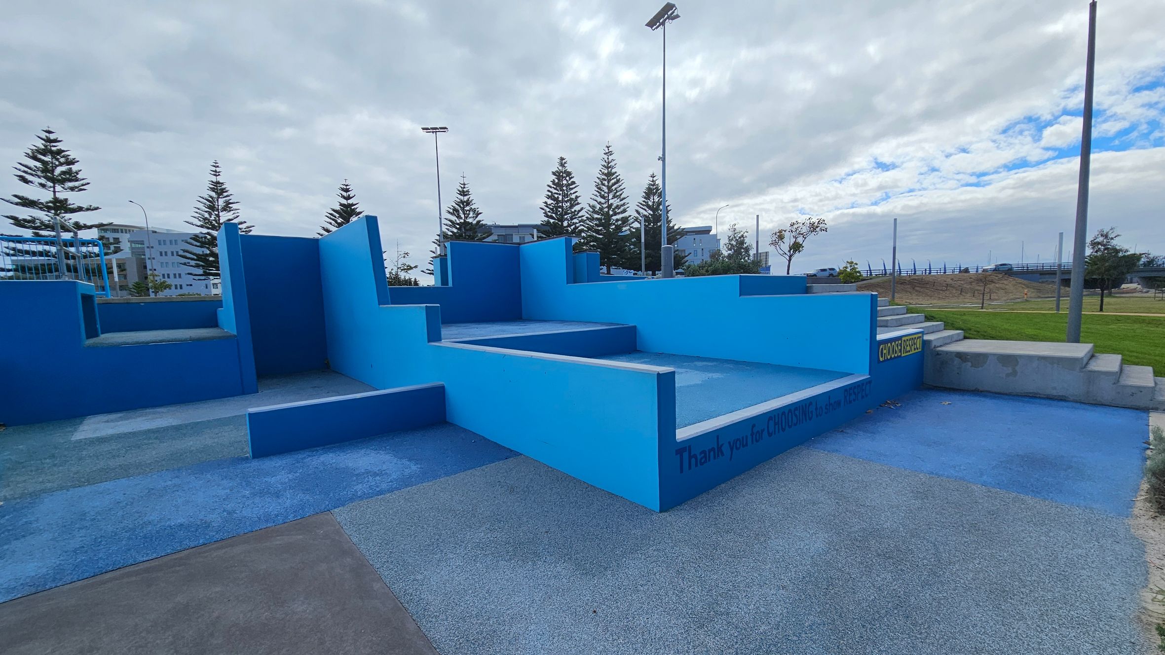 A geometric outdoor structure with various shades of blue, featuring multiple levels and steps. The ground is covered in textured, multicoloured surfaces. In the background, there are tall pine trees and a few buildings. A sign on the structure reads "Thank you for CHOOSING to show RESPECT." The area is open, with grassy patches and a few streetlights visible. A geometric outdoor structure with various shades of blue, featuring multiple levels and steps. The ground is covered in textured, multicoloured surfaces. In the background, there are tall pine trees and a few buildings. A sign on the structure reads "Thank you for CHOOSING to show RESPECT." The area is open, with grassy patches and a few streetlights visible.