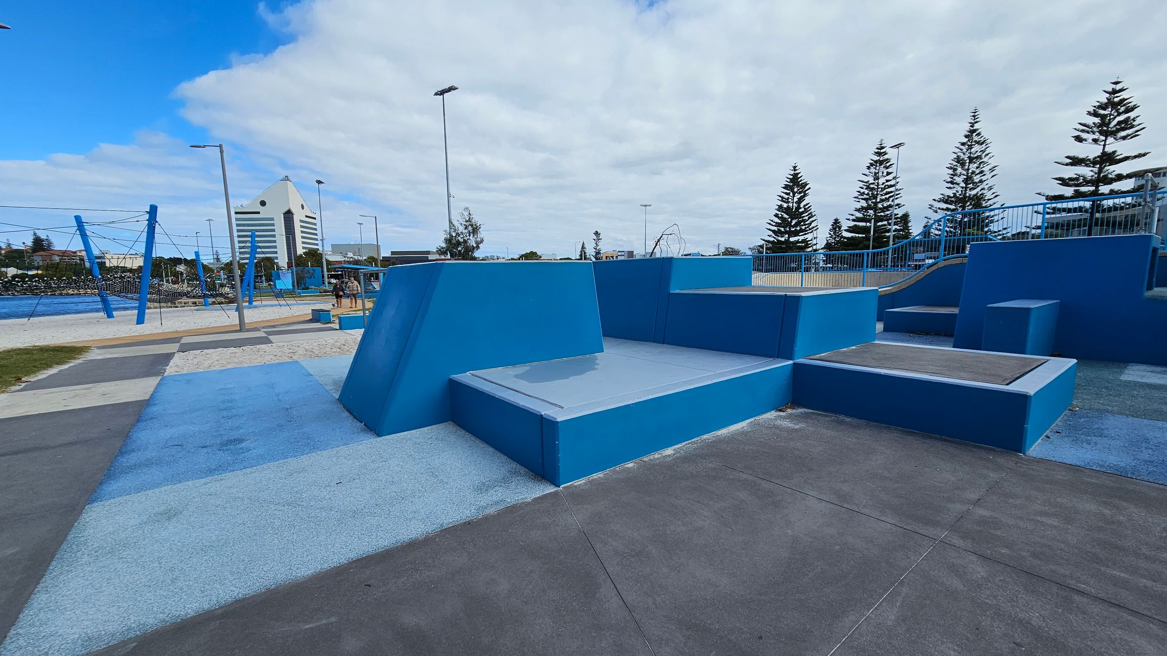 In the foreground, there are geometric structures in shades of blue and purple, resembling a skate park or playground. The ground is a mix of blue and purple surfaces. In the background, there are tall pine trees and a large, white, triangular-roofed building. To the left, there are blue poles and ropes, possibly part of a climbing structure, with a body of water visible beyond. The sky is overcast with patches of blue. In the foreground, there are geometric structures in shades of blue and purple, resembling a skate park or playground. The ground is a mix of blue and purple surfaces. In the background, there are tall pine trees and a large, white, triangular-roofed building. To the left, there are blue poles and ropes, possibly part of a climbing structure, with a body of water visible beyond. The sky is overcast with patches of blue.