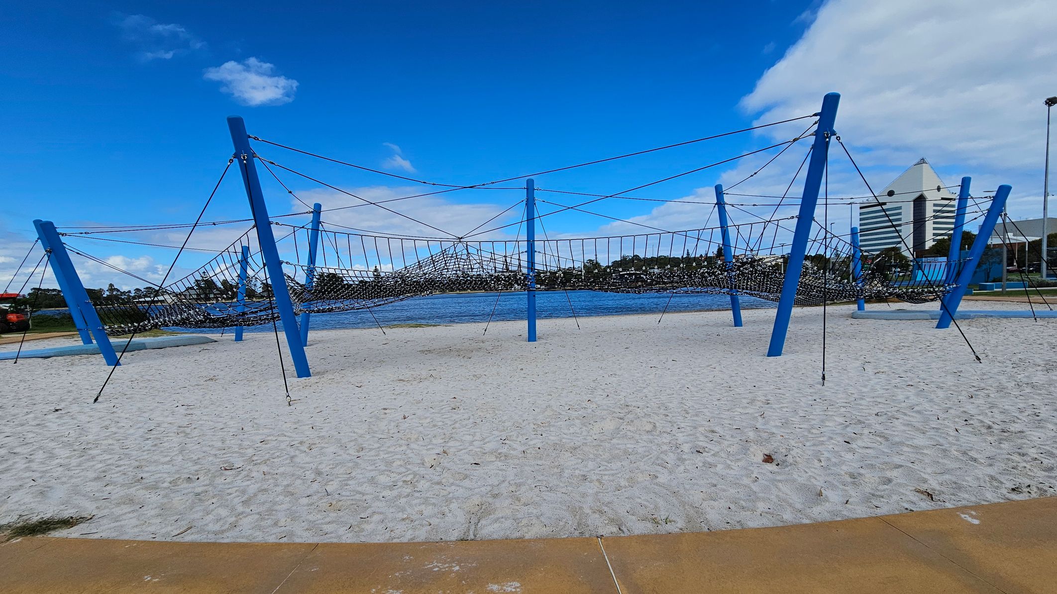 A playground structure with blue poles and a netted climbing area is set on a sandy surface. In the background, there is a body of water with trees lining the opposite shore. To the right, a tall, triangular-roofed building is visible. The foreground shows a paved area bordering the sand. A playground structure with blue poles and a netted climbing area is set on a sandy surface. In the background, there is a body of water with trees lining the opposite shore. To the right, a tall, triangular-roofed building is visible. The foreground shows a paved area bordering the sand.