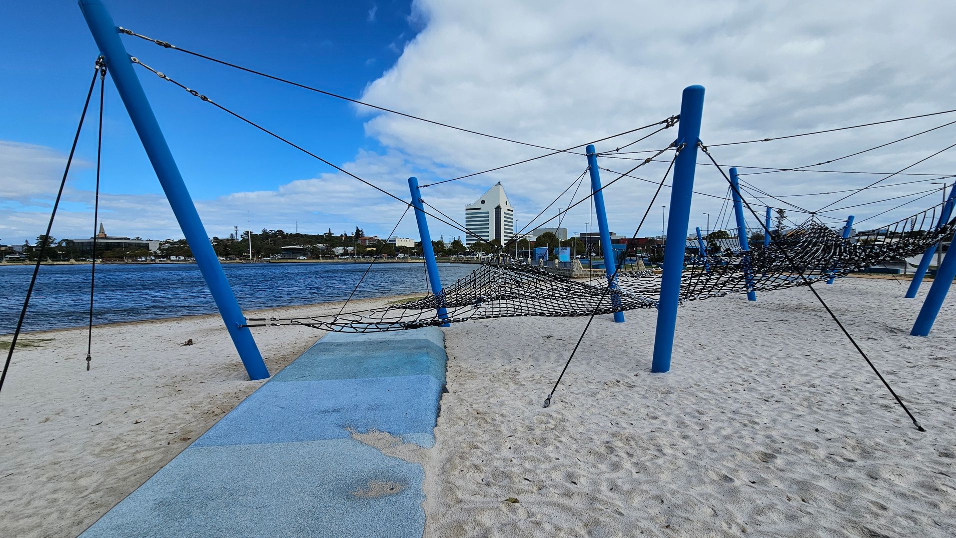 A sandy playground with blue poles supporting a net climbing structure. The foreground has a blue rubber matting area. In the background, a body of water is visible with a cityscape beyond, including a distinctive white triangular-roofed building. The sky is partly cloudy. A sandy playground with blue poles supporting a net climbing structure. The foreground has a blue rubber matting area. In the background, a body of water is visible with a cityscape beyond, including a distinctive white triangular-roofed building. The sky is partly cloudy.