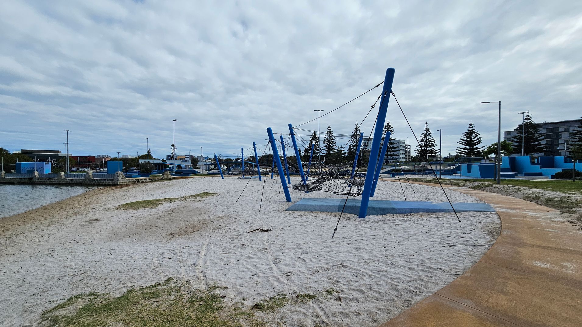 A sandy playground area with blue climbing structures and ropes is in the centre. On the left, there's a body of water bordered by a stone wall. In the background, there are several buildings and tall pine trees. A curved pathway runs along the right side, with grass patches scattered on the sand. Street lamps are visible throughout the area. A sandy playground area with blue climbing structures and ropes is in the centre. On the left, there's a body of water bordered by a stone wall. In the background, there are several buildings and tall pine trees. A curved pathway runs along the right side, with grass patches scattered on the sand. Street lamps are visible throughout the area.
