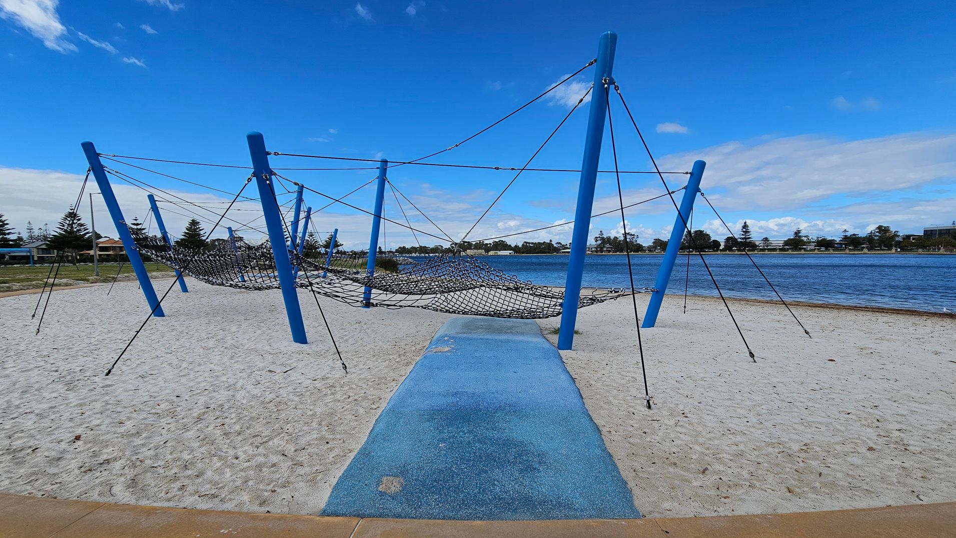 A playground structure with blue poles and a netted climbing area is set on a sandy surface. A blue pathway leads to the structure. In the background, there is a body of water with a shoreline lined with trees and buildings. The sky is clear with a few clouds. A playground structure with blue poles and a netted climbing area is set on a sandy surface. A blue pathway leads to the structure. In the background, there is a body of water with a shoreline lined with trees and buildings. The sky is clear with a few clouds.