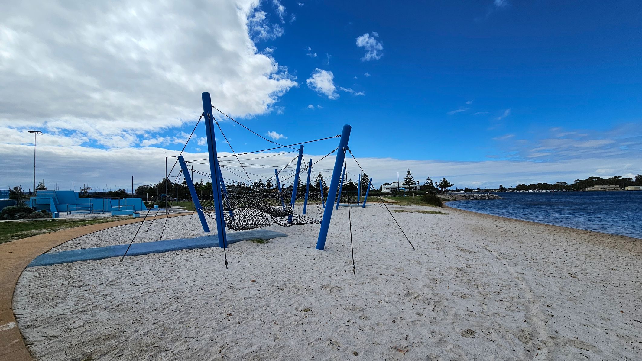 A sandy playground area with a large climbing structure made of blue poles and ropes is in the foreground. To the left, there are blue concrete structures and a pathway. In the background, there are trees and a body of water with a rocky shoreline. The sky is partly cloudy with patches of blue. A sandy playground area with a large climbing structure made of blue poles and ropes is in the foreground. To the left, there are blue concrete structures and a pathway. In the background, there are trees and a body of water with a rocky shoreline. The sky is partly cloudy with patches of blue.