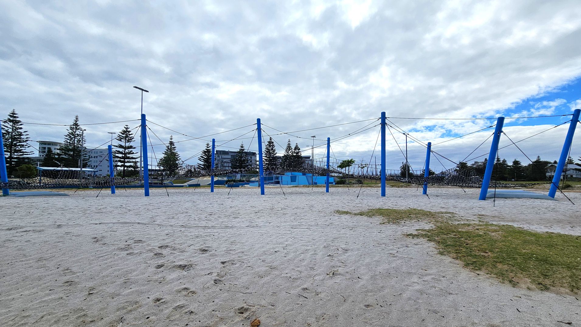 A sandy playground area with a large climbing structure made of blue poles and ropes. The structure is spread across the sand, with several poles connected by ropes forming a net-like design. In the background, there are trees and a few buildings visible. The ground has patches of grass mixed with the sand. The sky is overcast with some blue patches. A sandy playground area with a large climbing structure made of blue poles and ropes. The structure is spread across the sand, with several poles connected by ropes forming a net-like design. In the background, there are trees and a few buildings visible. The ground has patches of grass mixed with the sand. The sky is overcast with some blue patches.
