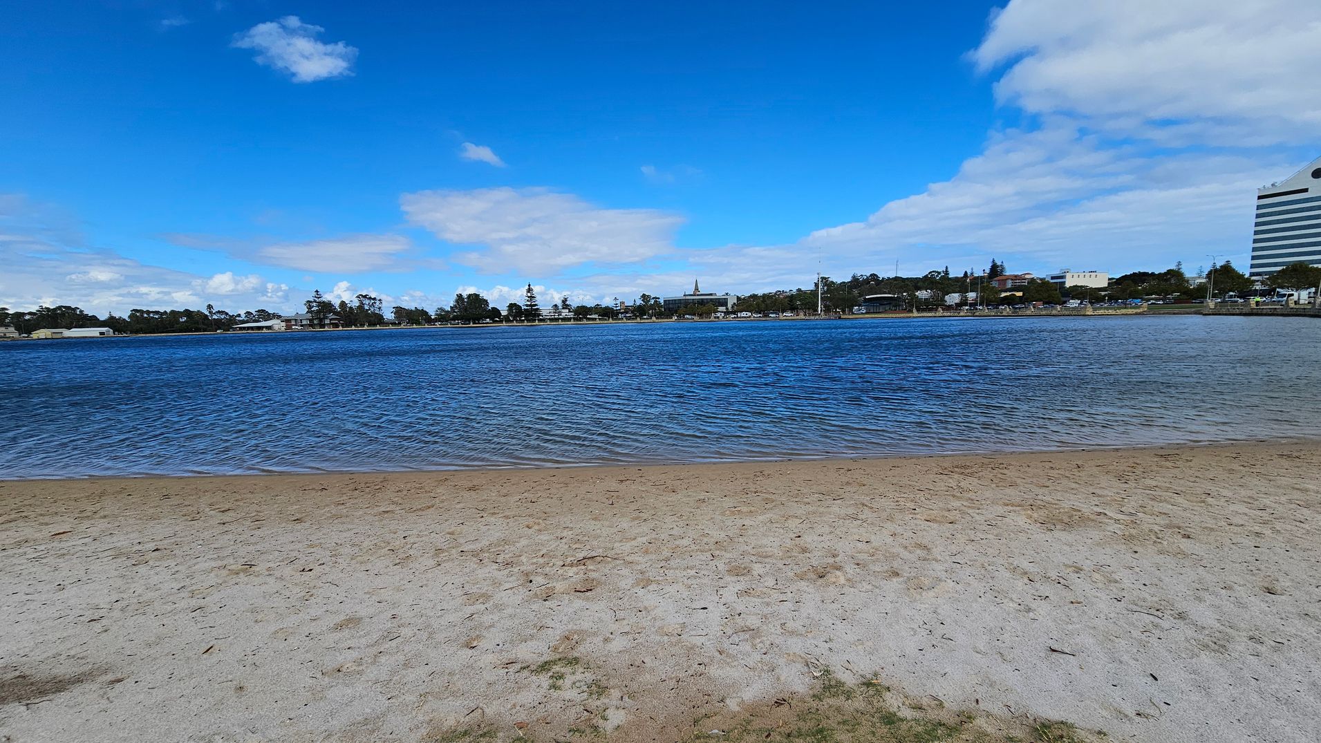 A sandy beach in the foreground leads to a body of water with gentle ripples. Across the water, a distant shoreline features a mix of trees and buildings, including a tall structure on the right. The sky is mostly clear with a few scattered clouds. A sandy beach in the foreground leads to a body of water with gentle ripples. Across the water, a distant shoreline features a mix of trees and buildings, including a tall structure on the right. The sky is mostly clear with a few scattered clouds.