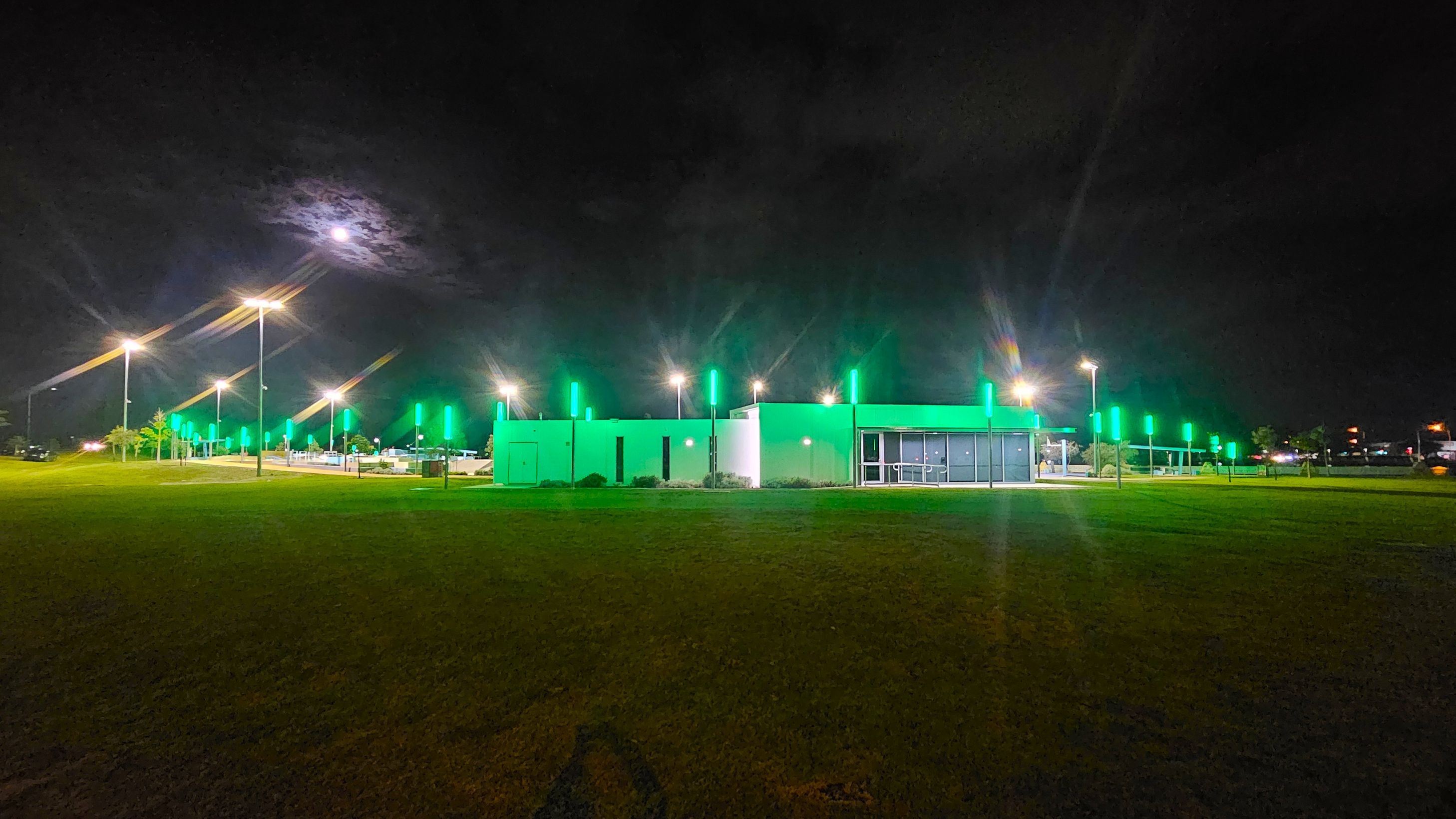 A modern building is centrally located, illuminated with green lights. To the left, a row of street lamps lines a pathway, casting a warm glow. The foreground features a grassy area, while the background is a dark night sky with a bright moon partially obscured by clouds. The right side of the building has large glass windows, and there are more green-lit poles extending into the distance. A modern building is centrally located, illuminated with green lights. To the left, a row of street lamps lines a pathway, casting a warm glow. The foreground features a grassy area, while the background is a dark night sky with a bright moon partially obscured by clouds. The right side of the building has large glass windows, and there are more green-lit poles extending into the distance.
